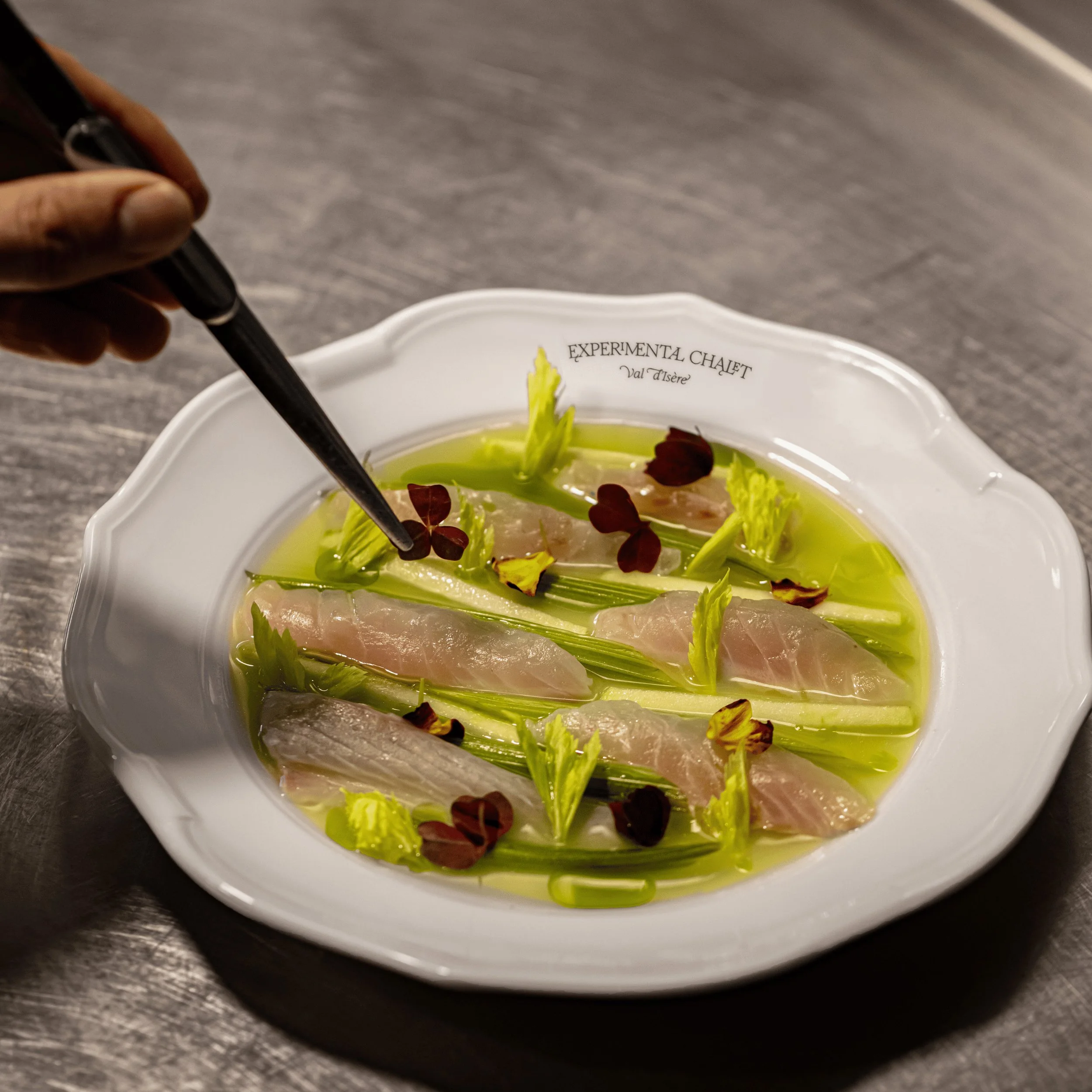 Colocando manualmente pequenas folhas vermelhas com uma pinça em um prato de peixe, aipo e molho verde.