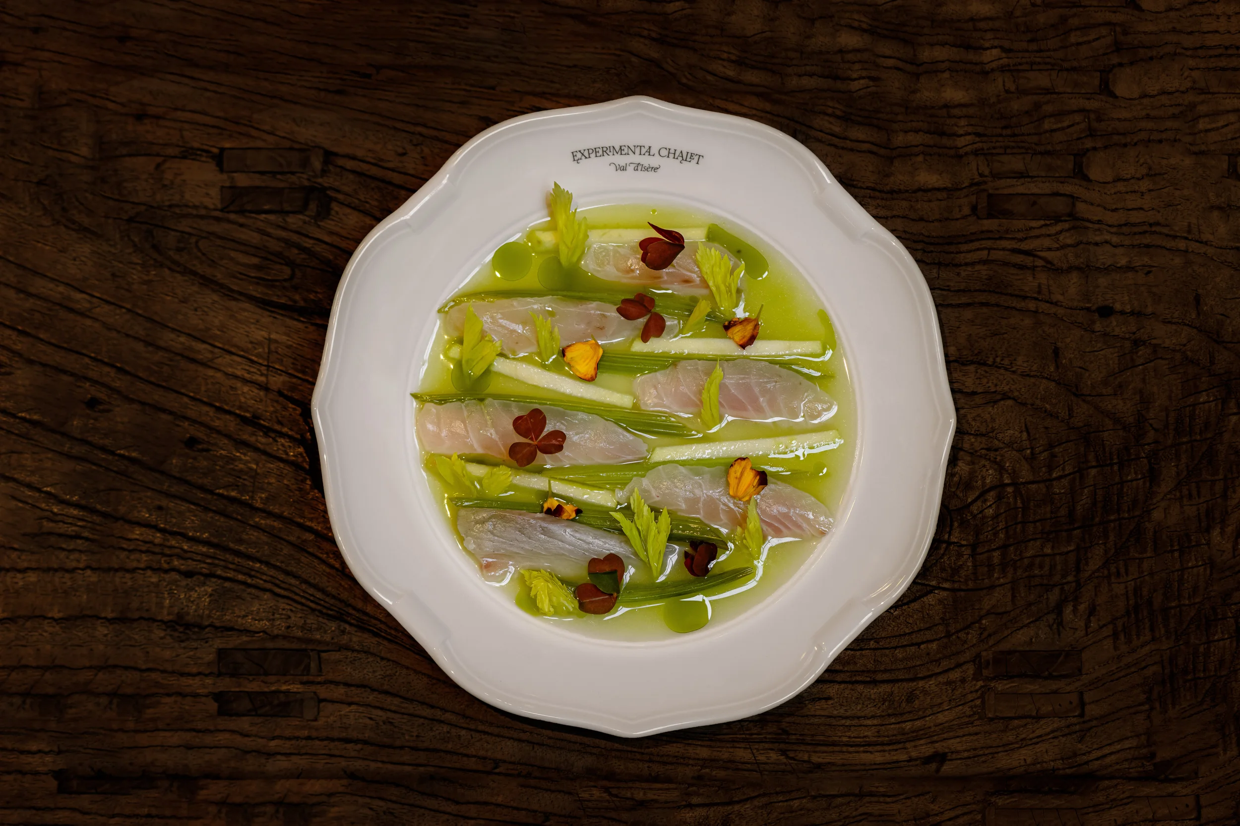 Prato branco com fatias de peixe cru, molho verde, folhas de aipo, palitos de maçã e pétalas de flores comestíveis na mesa de madeira.