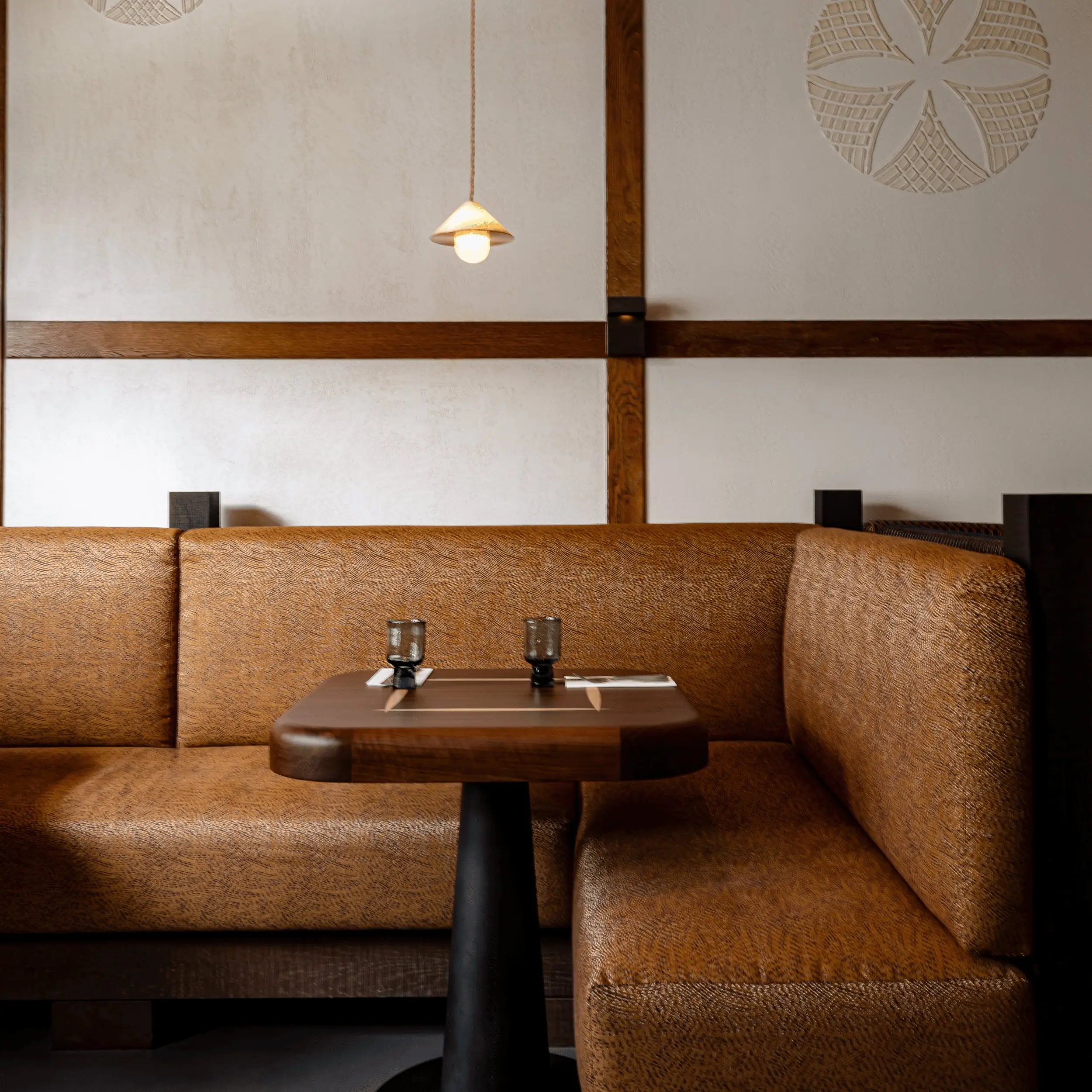 Estande de restaurante aconchegante com assentos texturizados marrons, mesa de madeira, copos e uma luz suspensa acima.