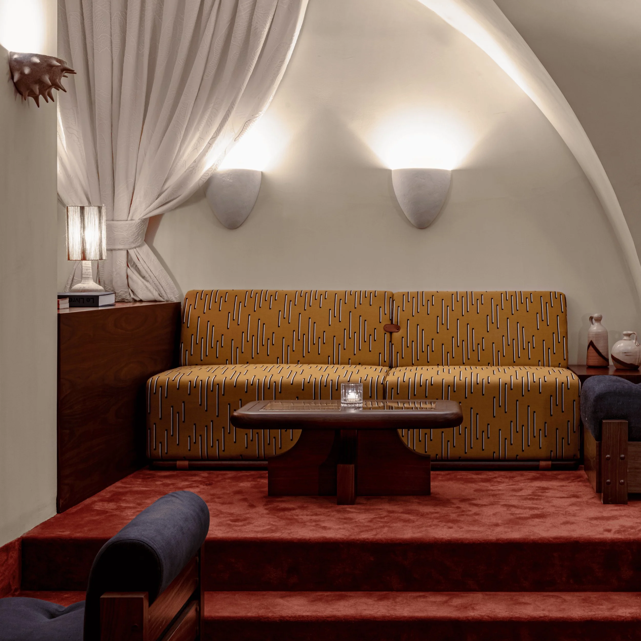 Sala de estar aconchegante com sofá de mostarda estampado, cadeiras escuras, mesa de madeira e apliques suaves.