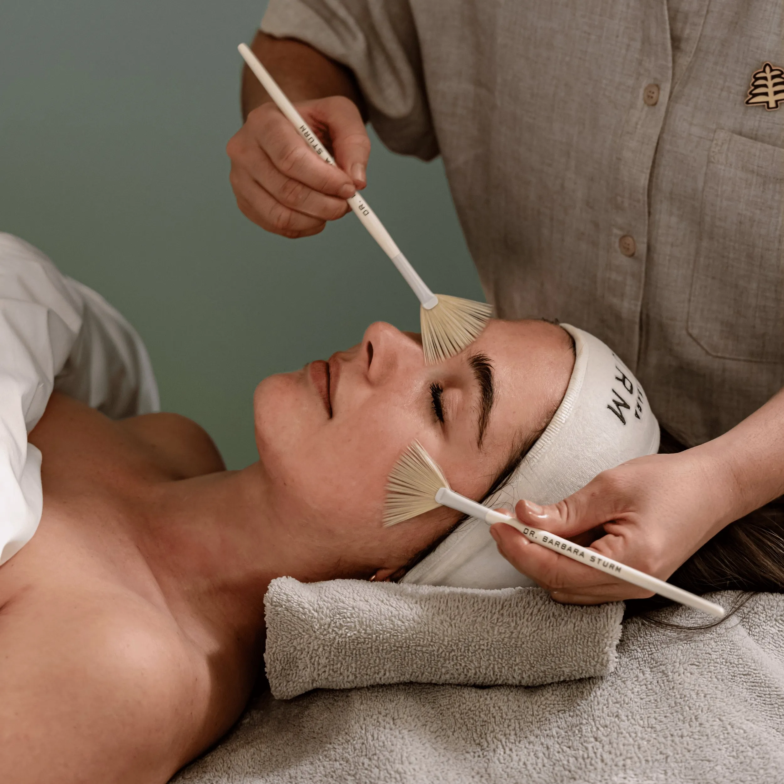 Pessoa recebendo tratamento facial com escovas na testa e bochecha, deitada na cama coberta com toalha.