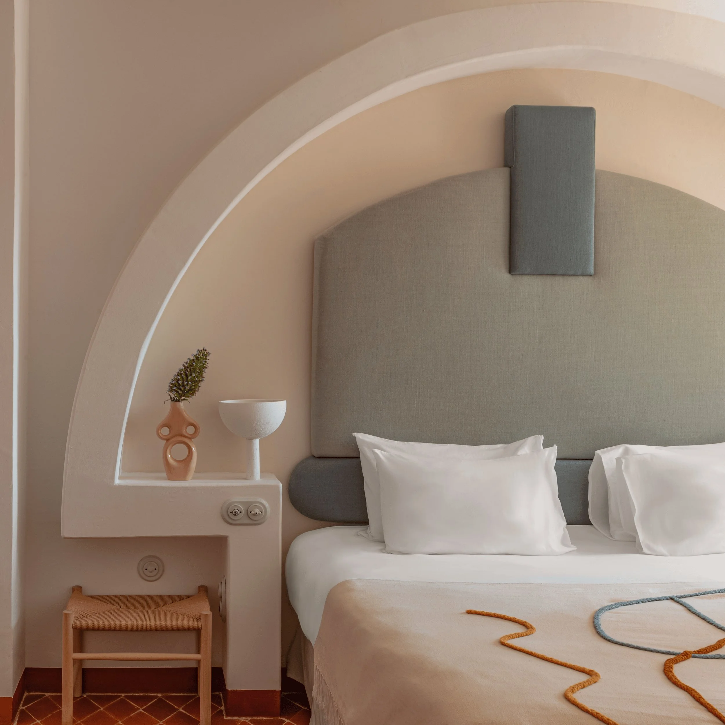 Habitación de hotel minimalista con una pared arqueada de color beige, cabecera tapizada en gris, almohadas blancas y taburete de madera.