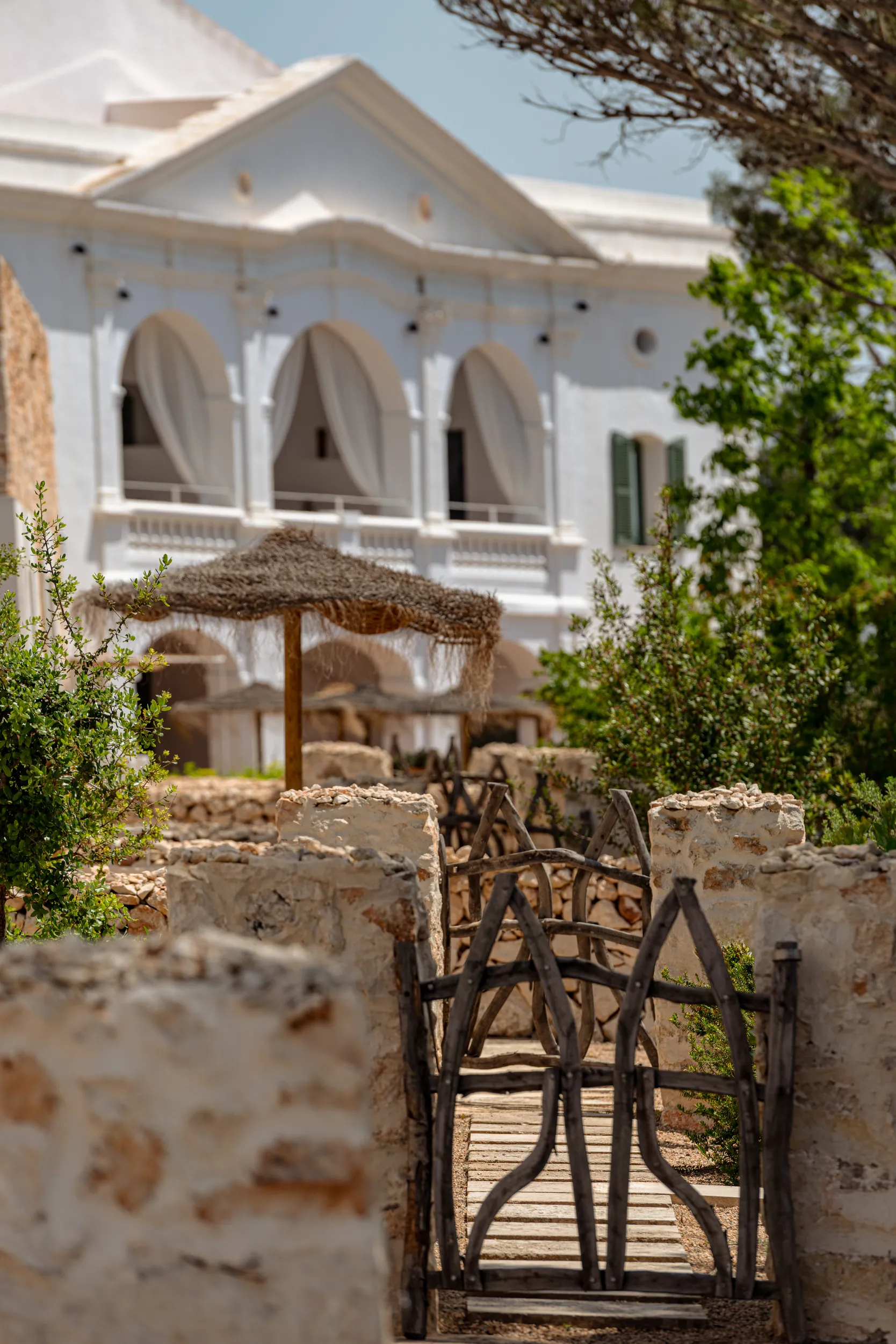 Rustic stone path with wooden gates leading to white building with arched windows and thatched umbrellas.