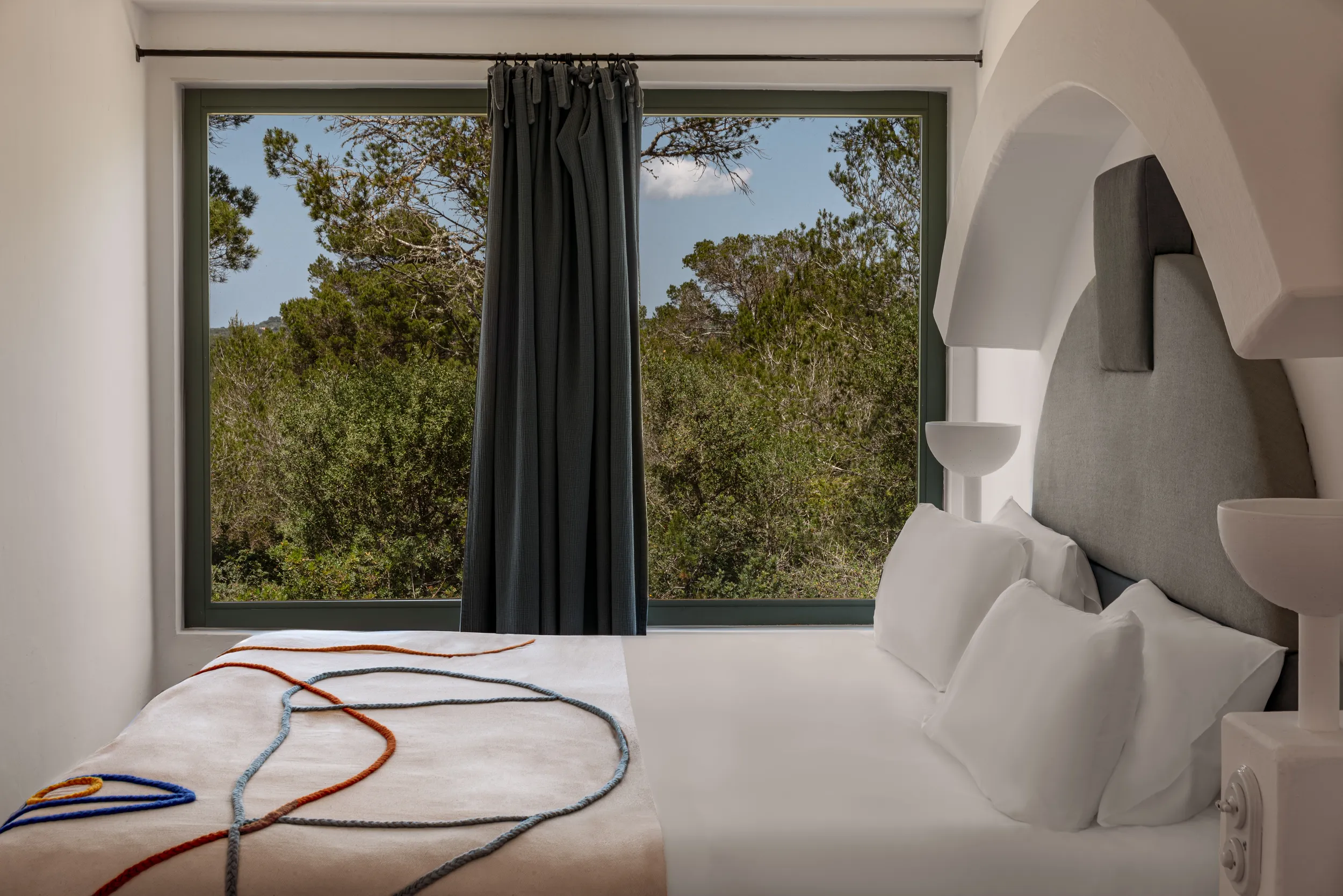Cama moderna con sábanas blancas y colorida manta frente a una ventana grande con vista al bosque verde.