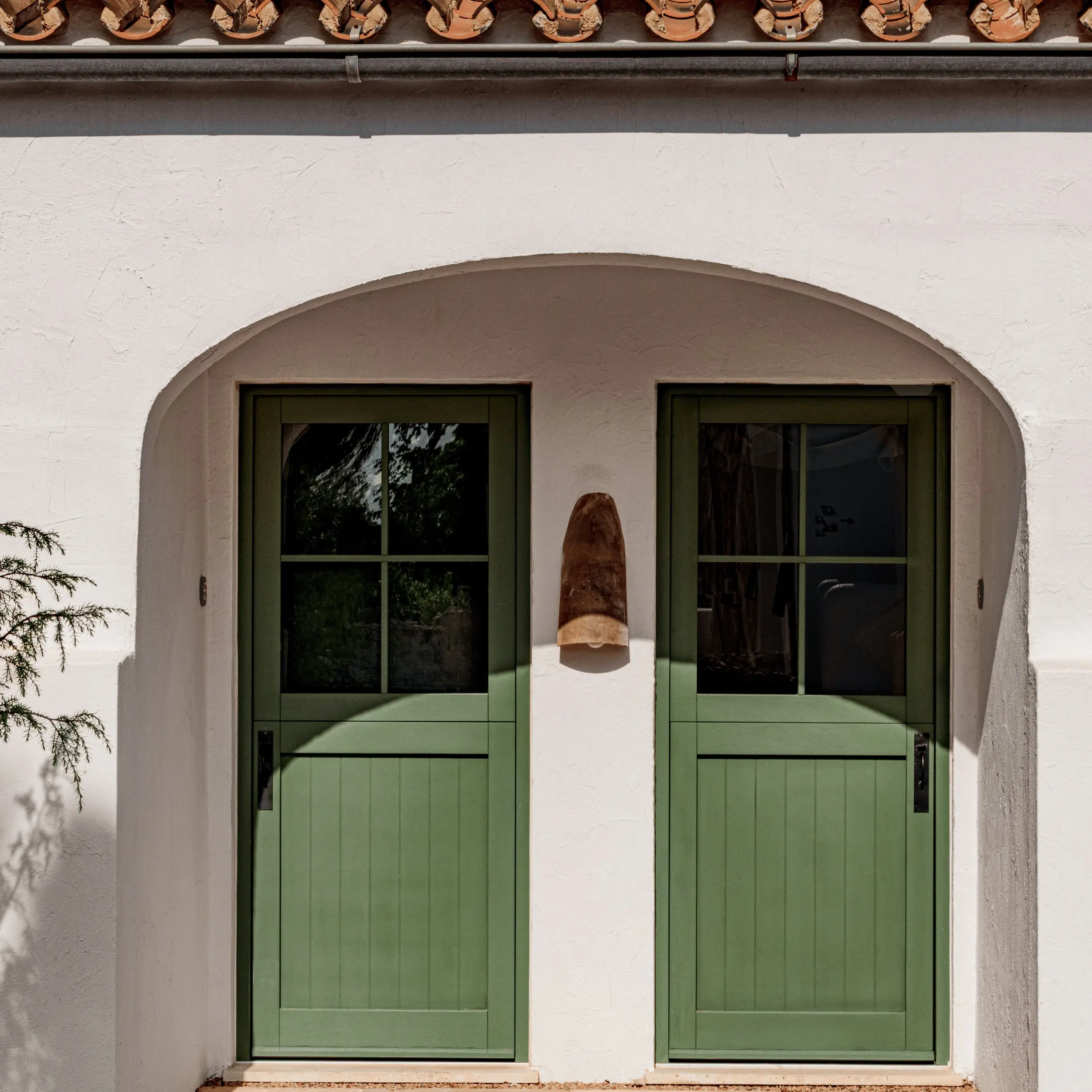 Dos puertas de madera verde con paneles de vidrio bajo un arco de estuco blanco con una lámpara rústica entre ellas.