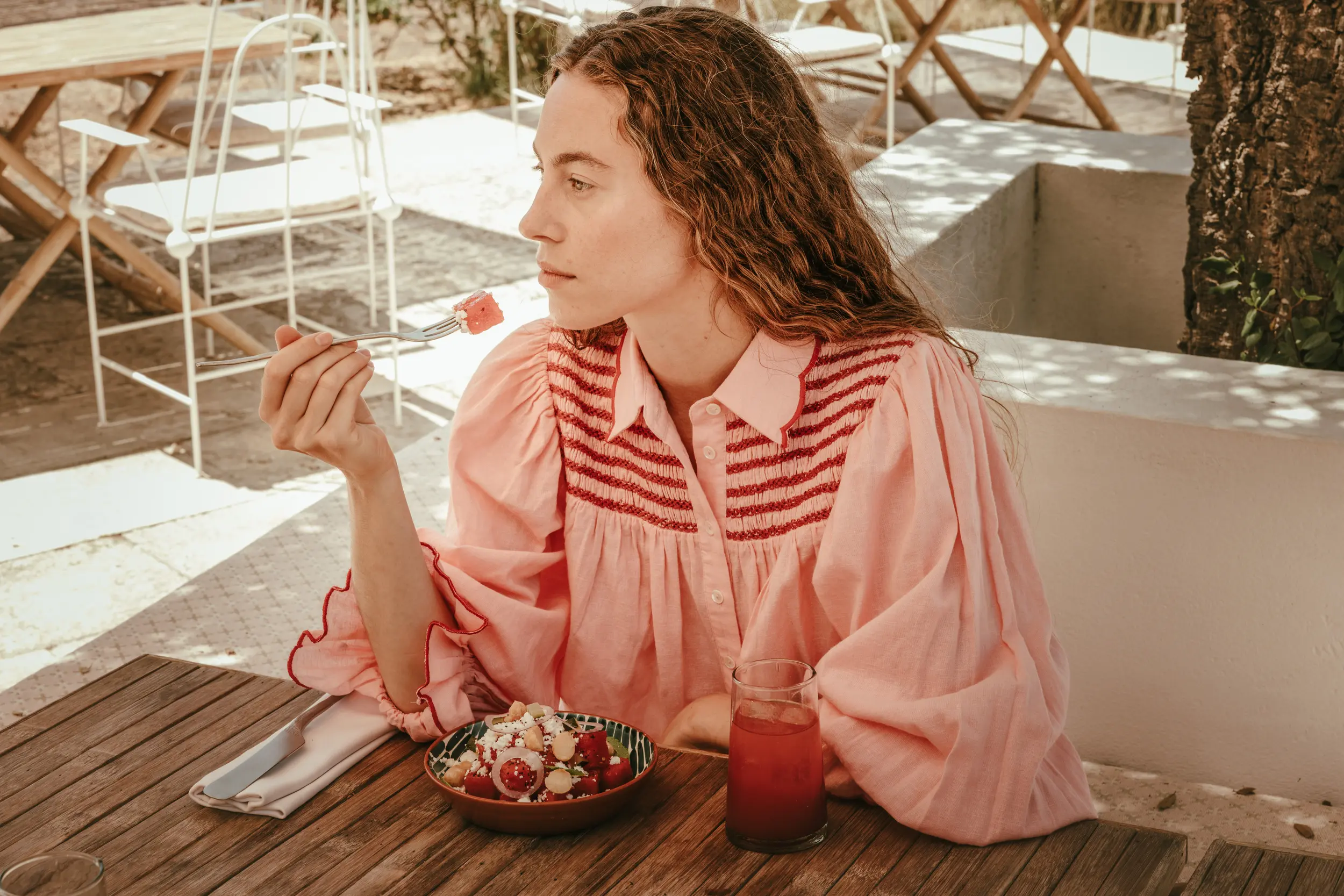 Mujer en blusa rosa comiendo ensalada de frutas en una mesa de madera al aire libre con un vaso de jugo.
