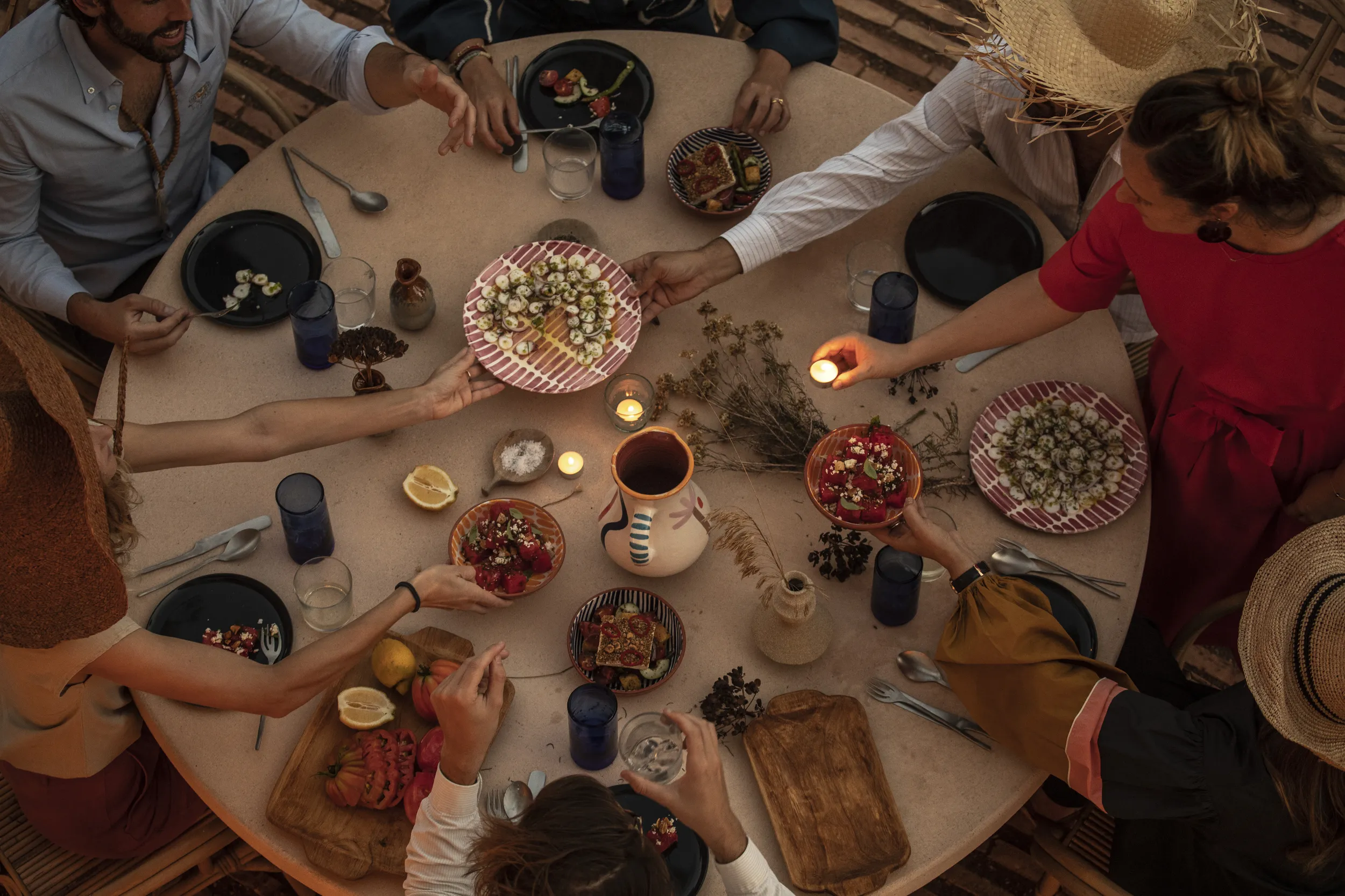 Vista aérea de seis personas compartiendo comida y encendiendo velas alrededor de una mesa redonda al aire libre.