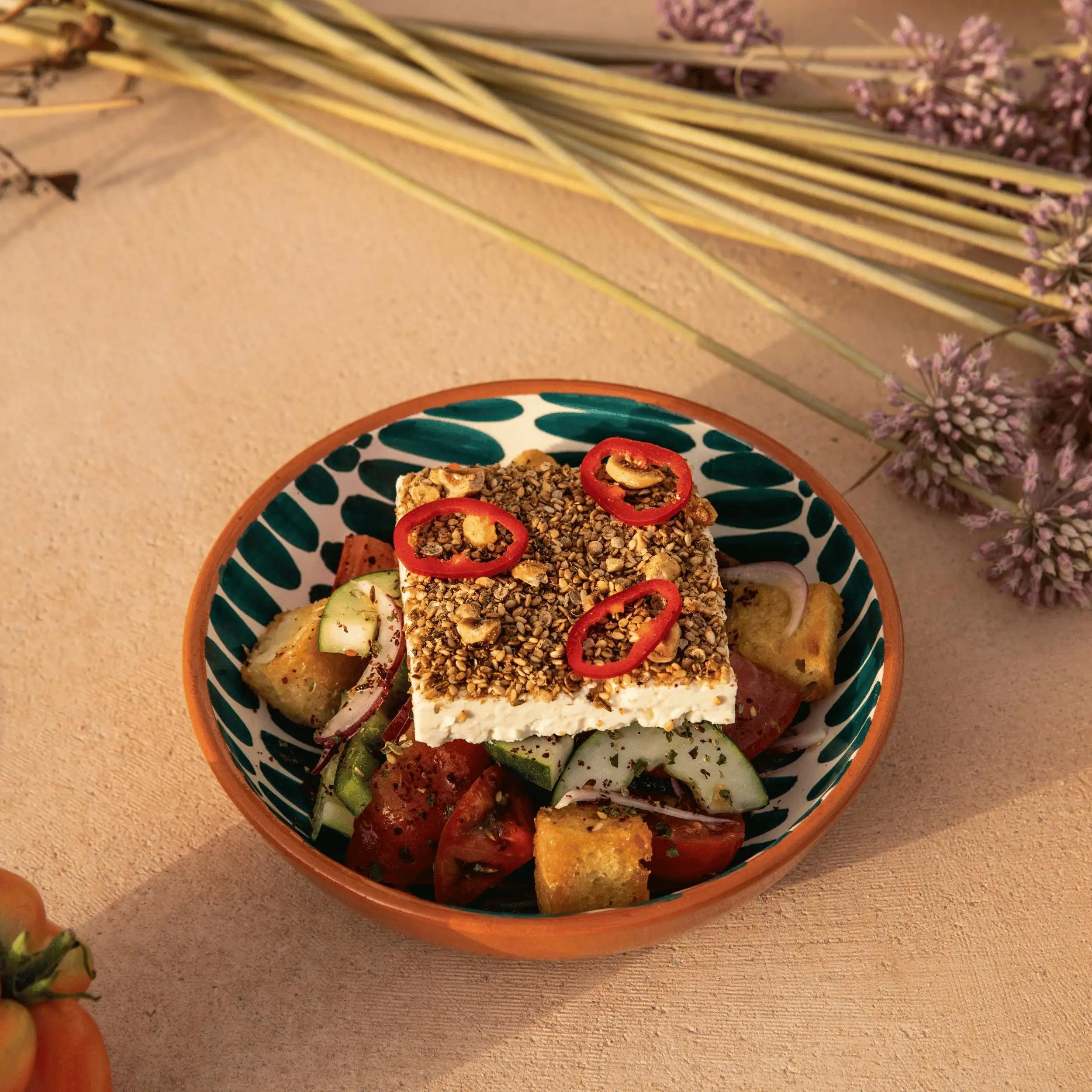 Tazón con queso feta cubierto con semillas y chile, sobre una ensalada de tomate, pepino, cebolla y picatostes.