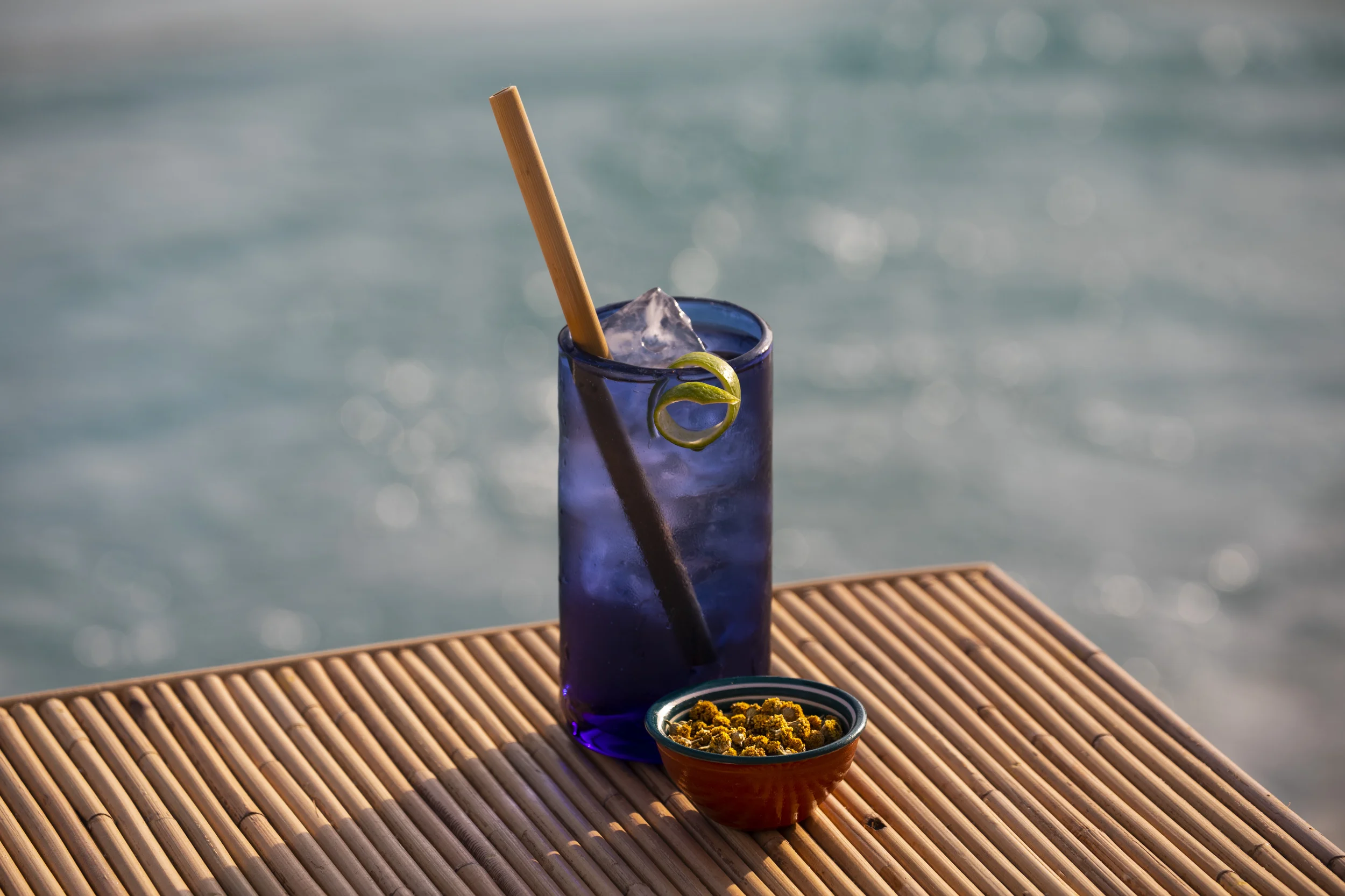Un vaso azul con hielo, paja de bambú y lima se retuerce junto a un tazón pequeño sobre una superficie de madera junto al agua.