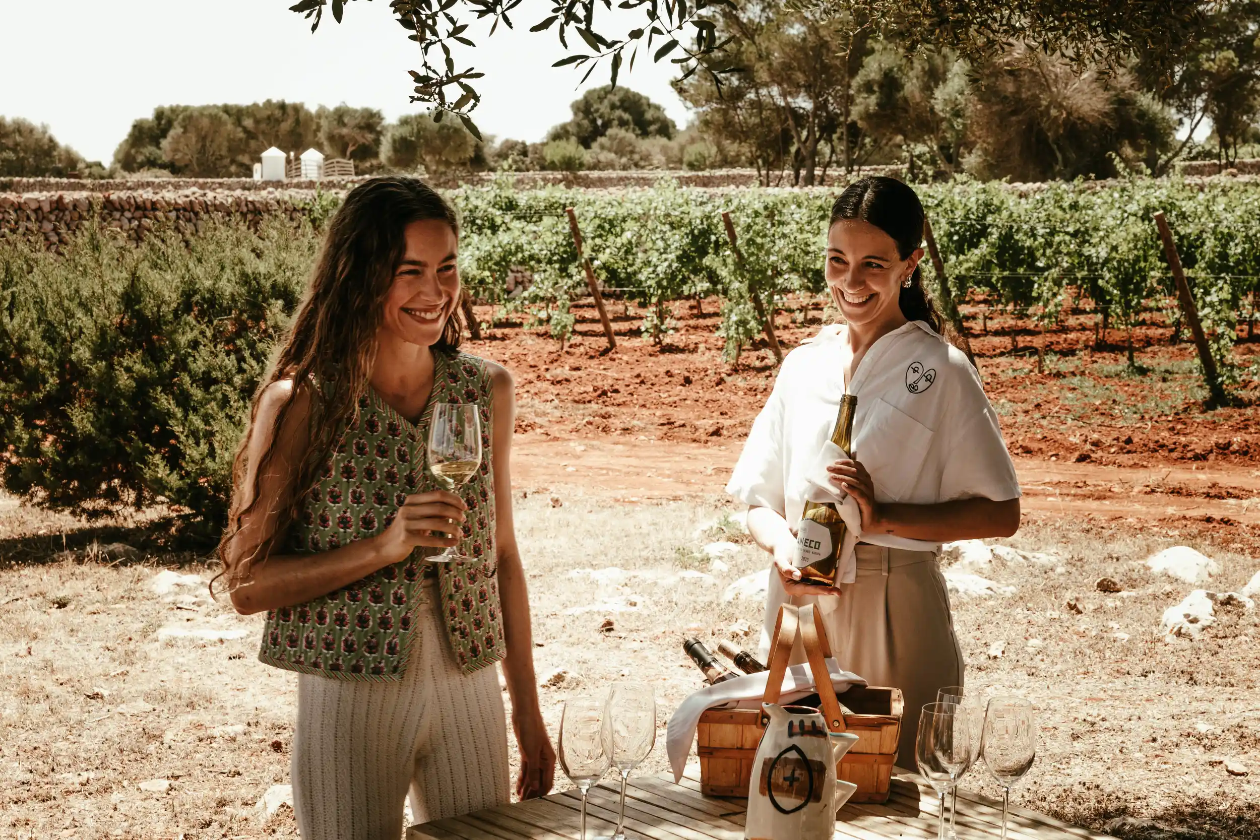 Two women smiling outdoors near vineyard, one holding wine glass, other holding wine bottle by table.