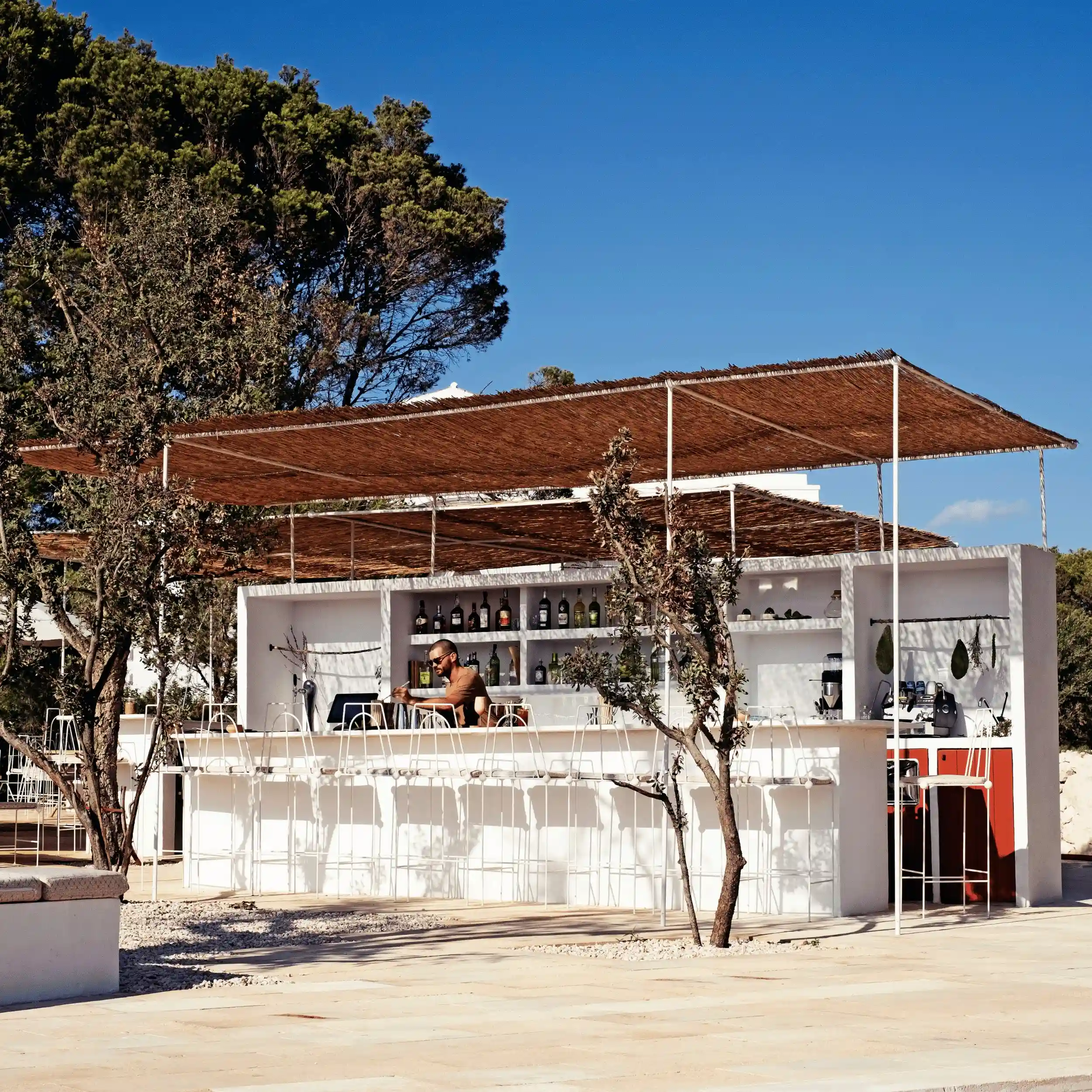 Bar blanco al aire libre con taburetes altos y un camarero que prepara bebidas bajo sombrillas tejidas.