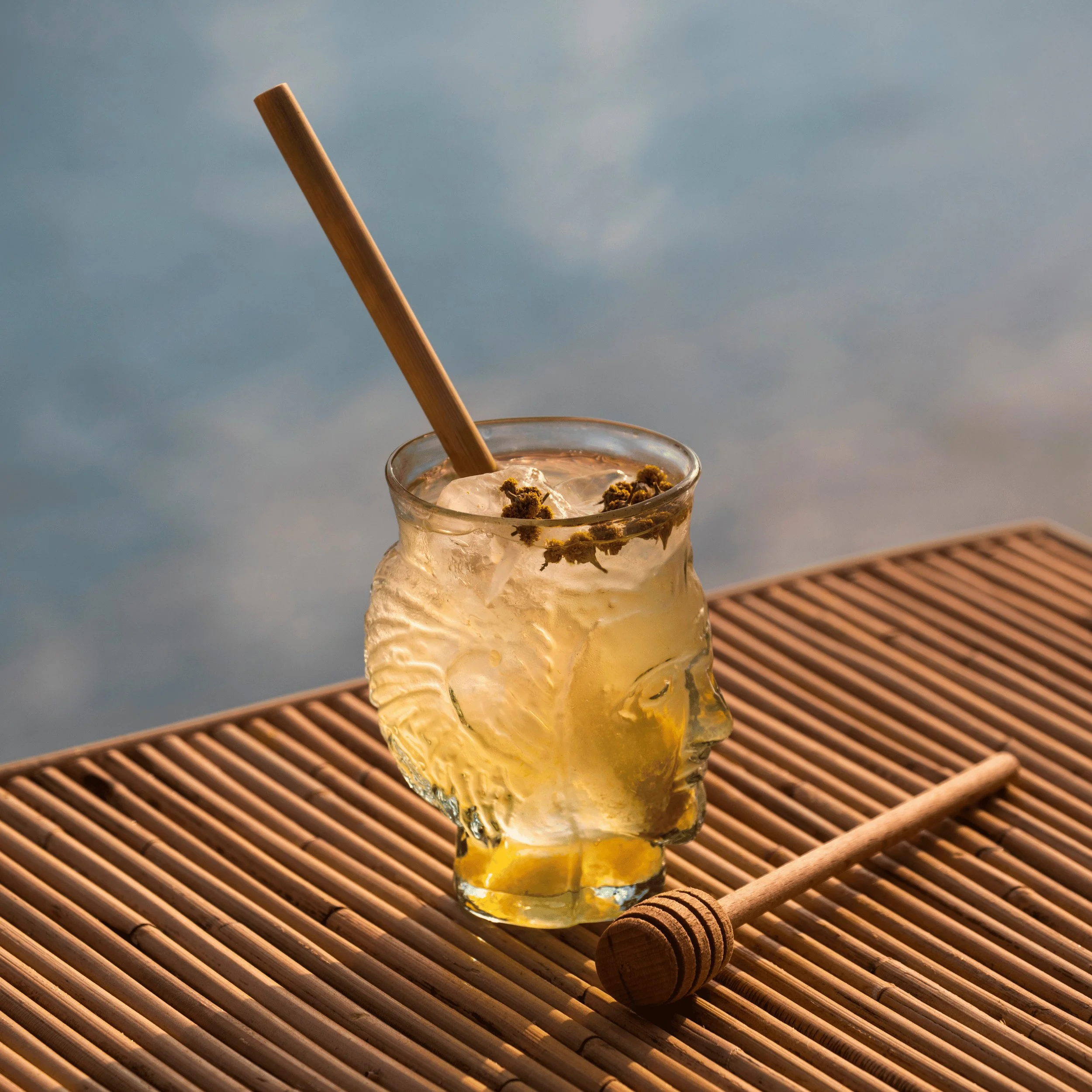 Vaso con forma de cabeza humana con cóctel amarillo helado y agitador de madera sobre superficie de bambú.