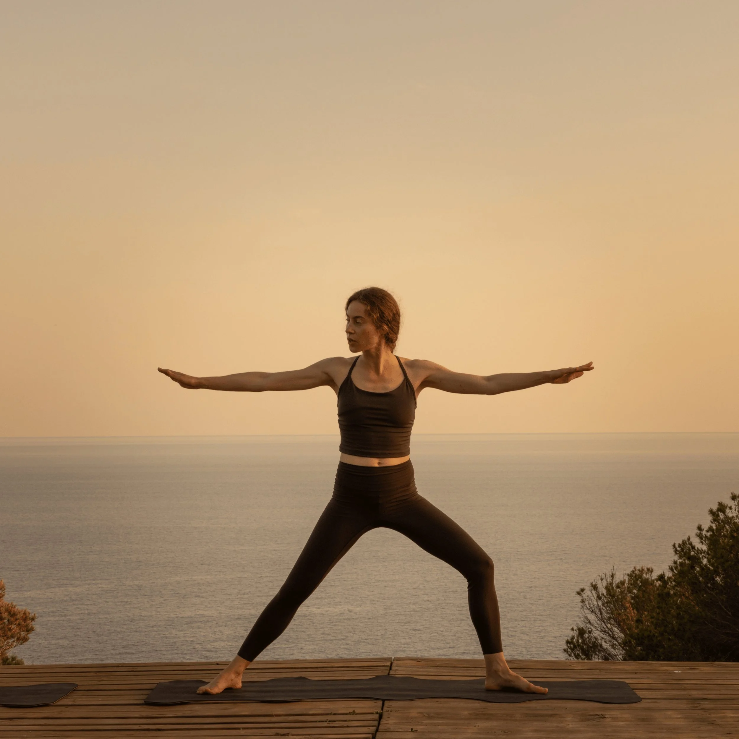 Mujer en ropa deportiva negra haciendo yoga en postura de guerrera en cubierta con vistas al mar en calma al atardecer.