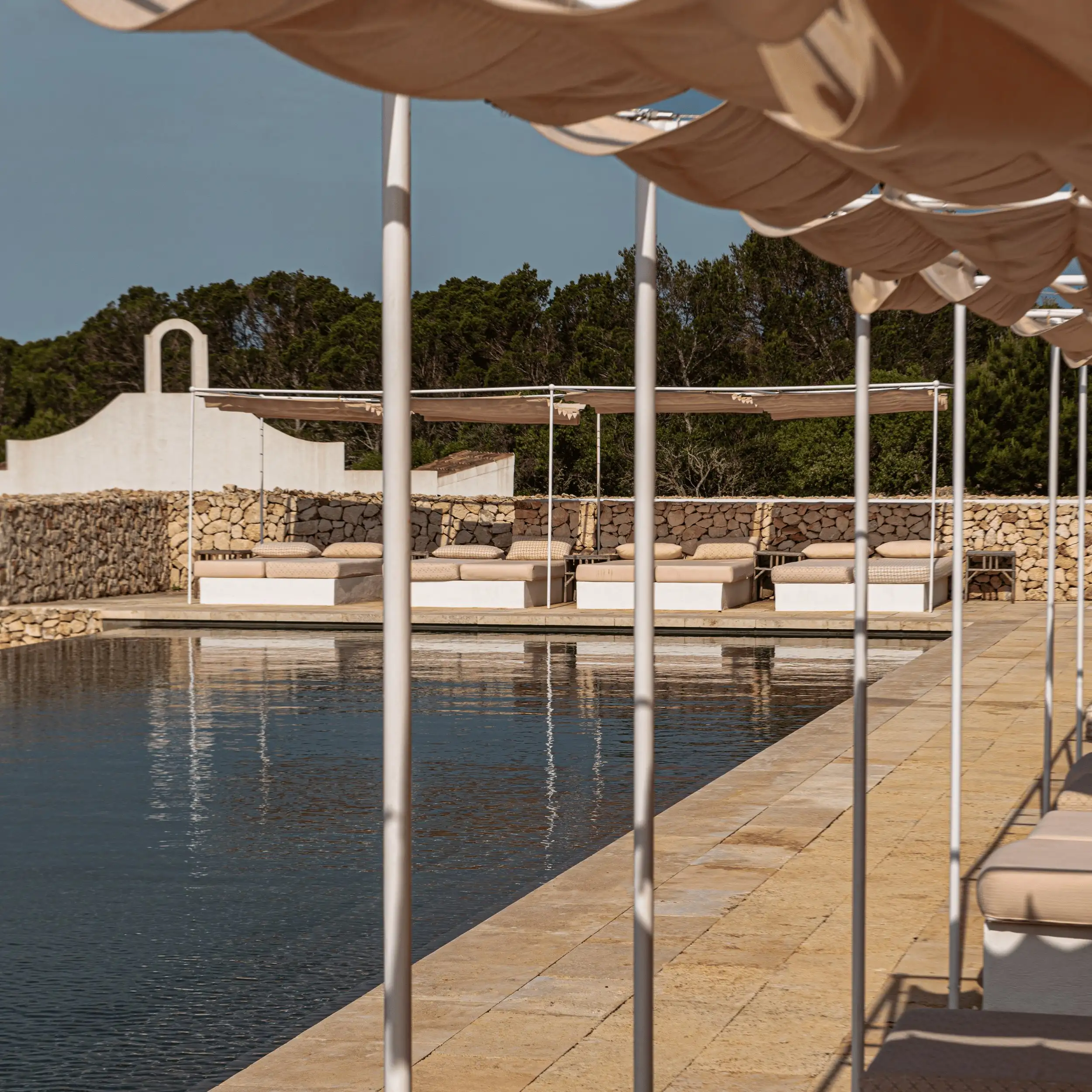 Piscina al aire libre con tumbonas cubiertas con toldos de color beige y paredes de piedra bajo un cielo despejado.
