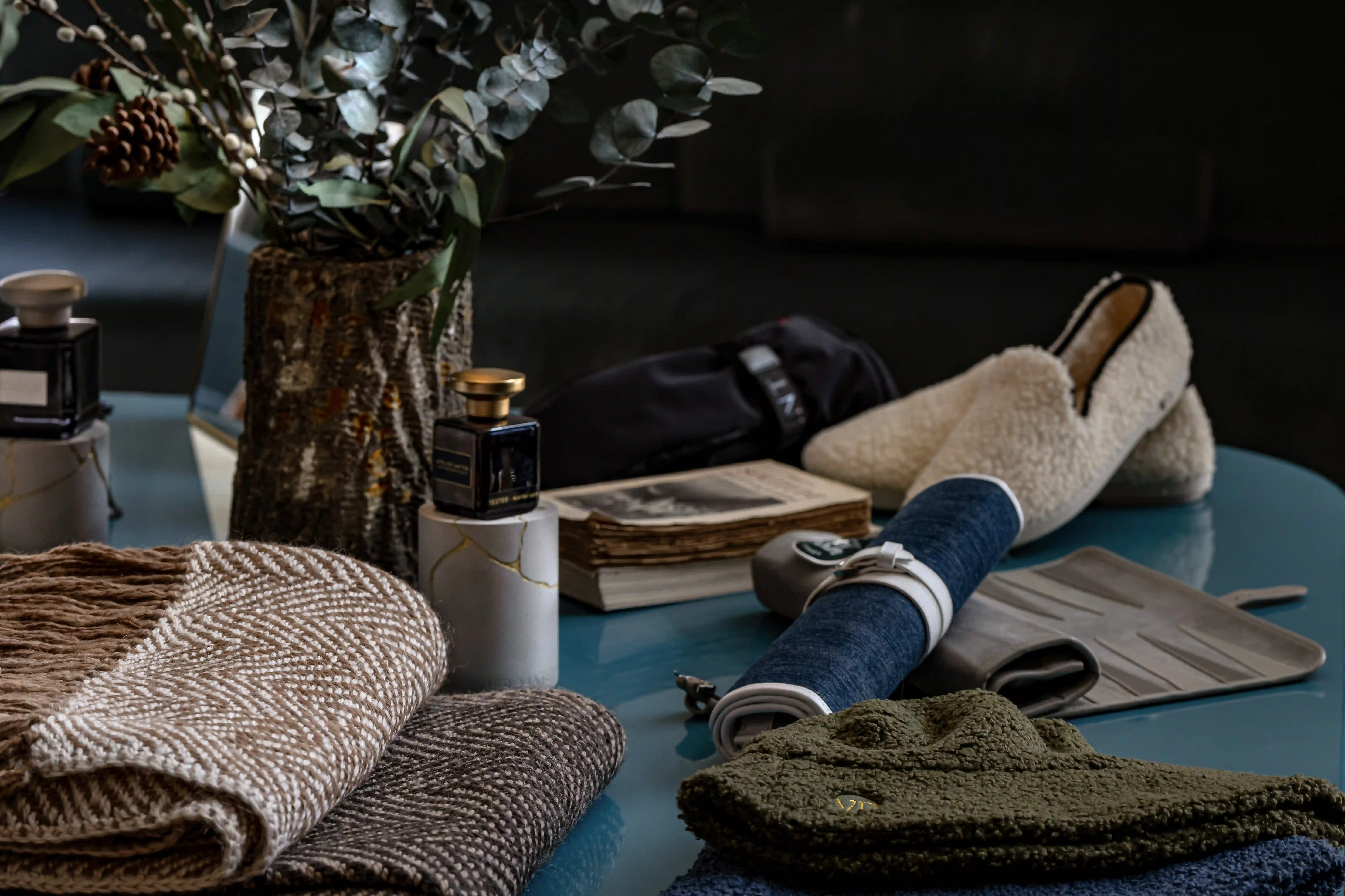 Cozy home items on table: knit blankets, sheepskin slippers, rolled mat, vintage book, scented bottles, and plant vase.