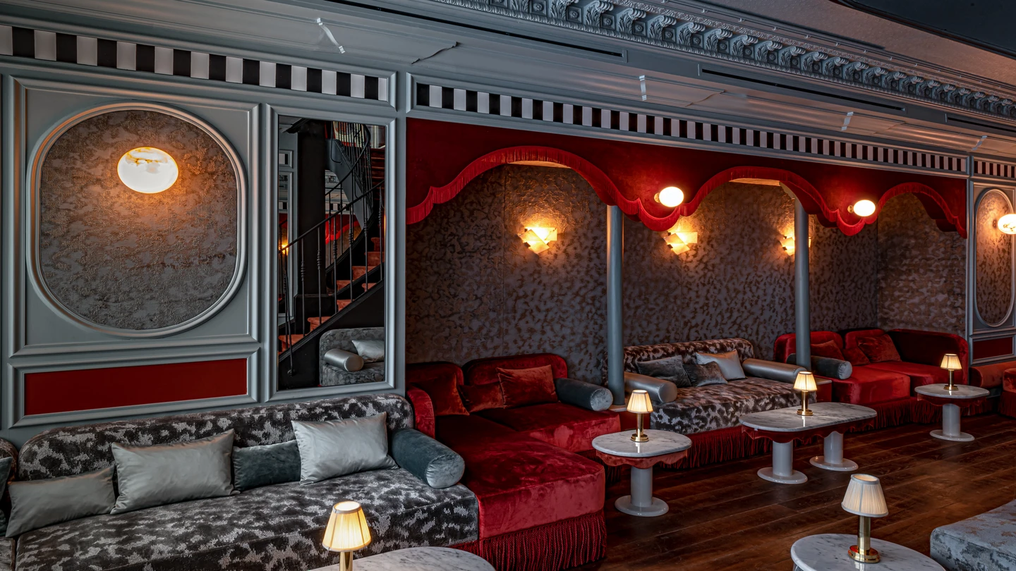 Lounge luxuoso com sofás estampados, mesas pequenas, luminárias macias e paredes cobertas de veludo vermelho.