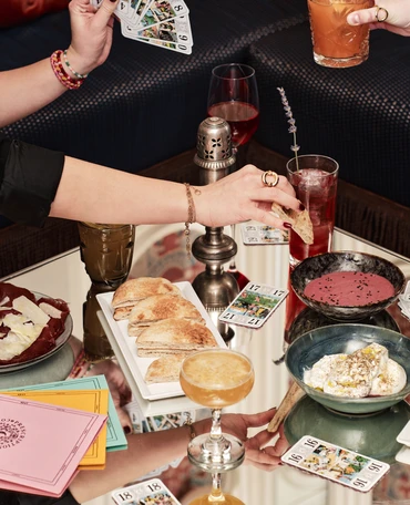 Pessoas desfrutando de bebidas, lanches e cartas de tarô em uma mesa de vidro refletivo.