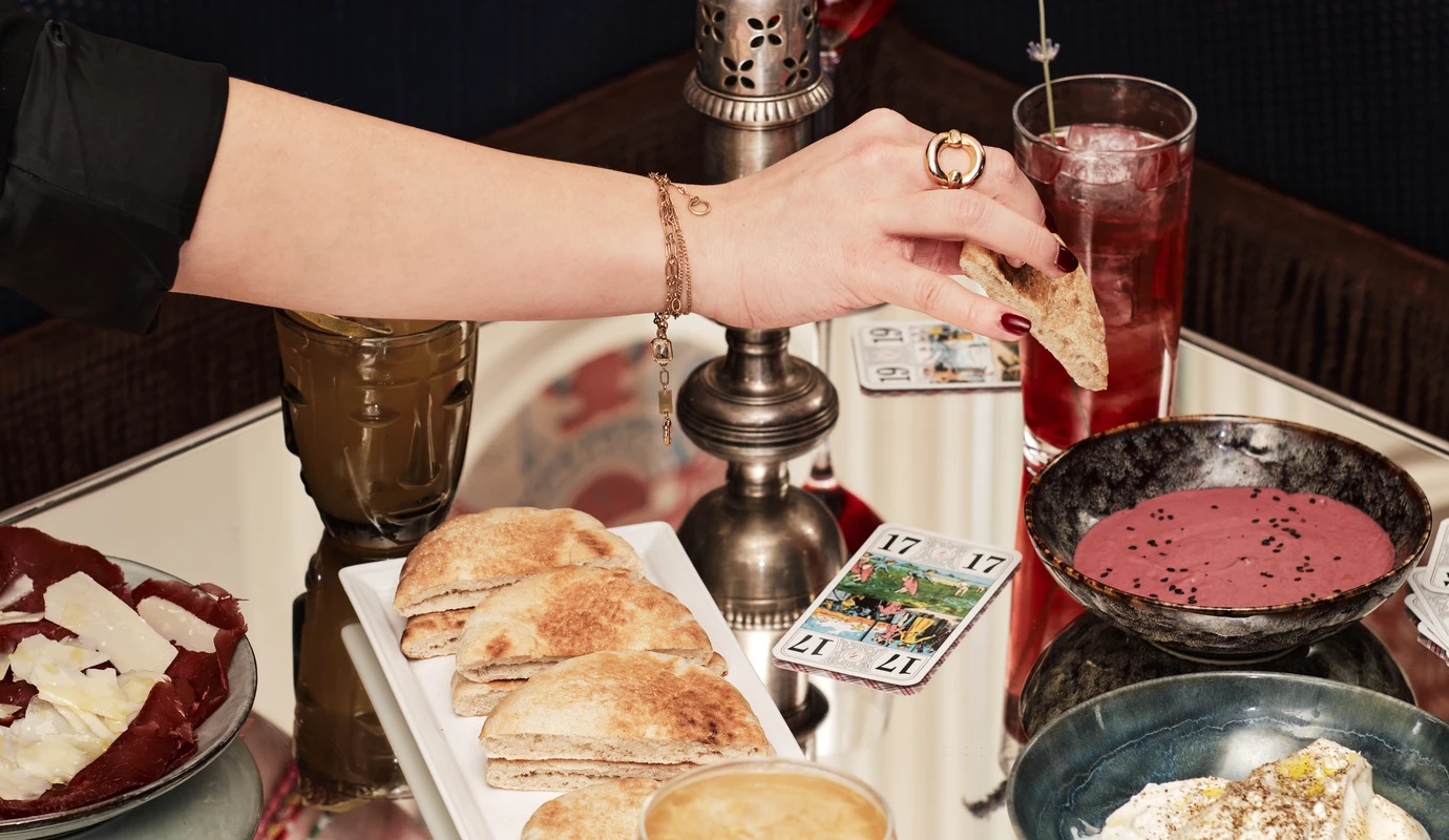 Mergulhando pão achatado à mão em uma tigela de homus rosa em uma mesa espelhada com alimentos e bebidas.