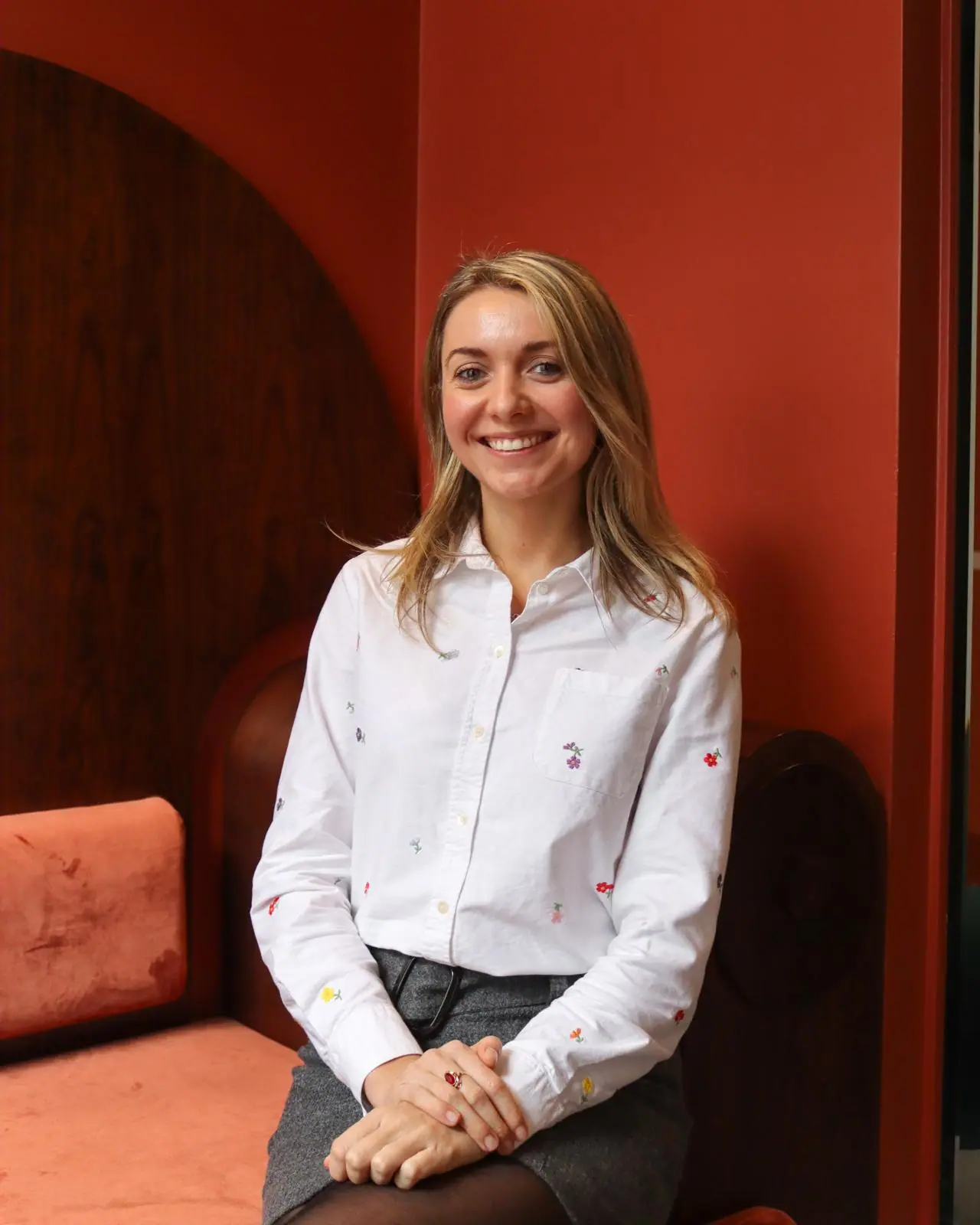 Smiling woman in a white shirt with small floral patterns sits on an orange bench against a red wall.
