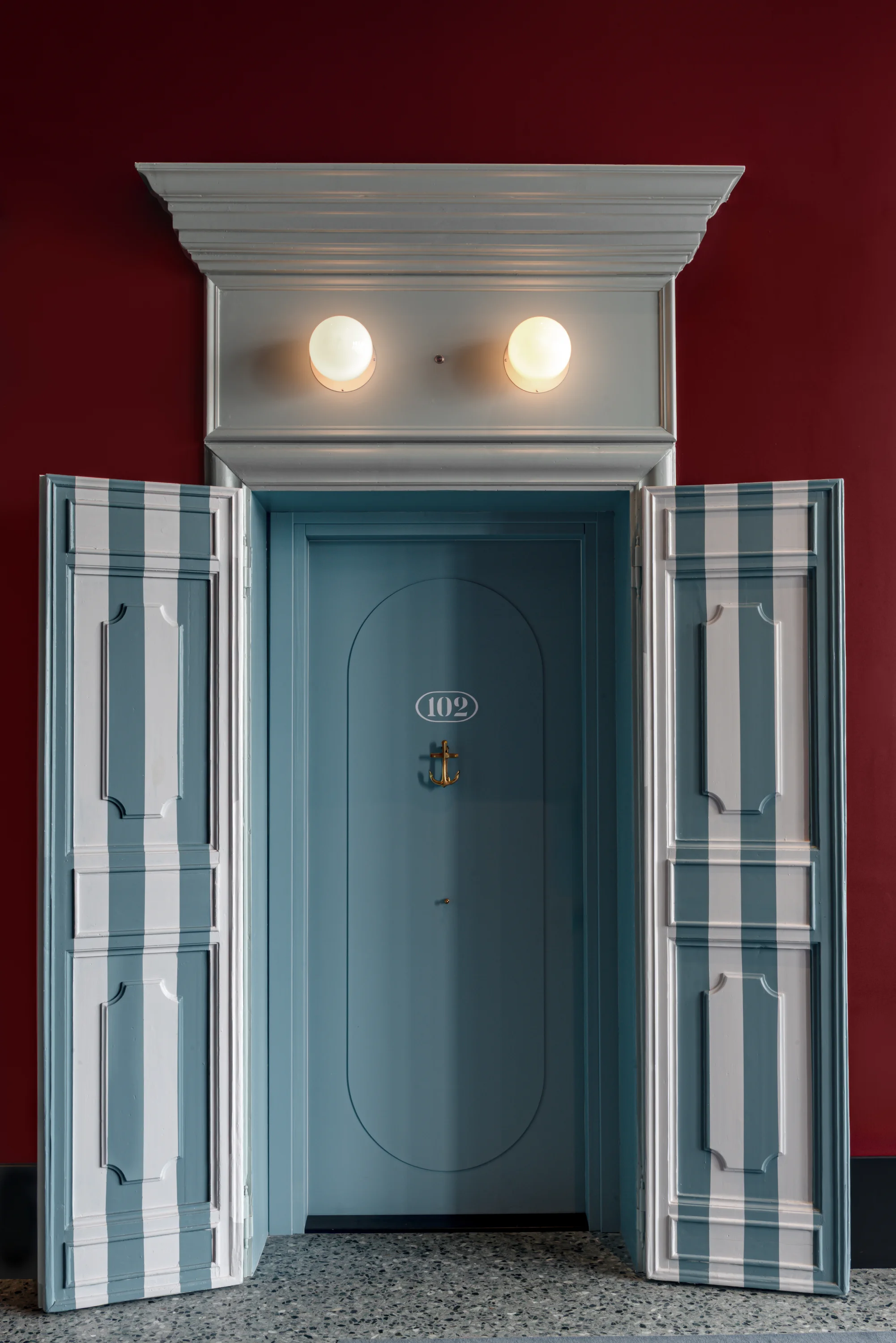 Puerta azul numerada 102 con manija en forma de ancla y persianas a rayas a juego en la pared roja.