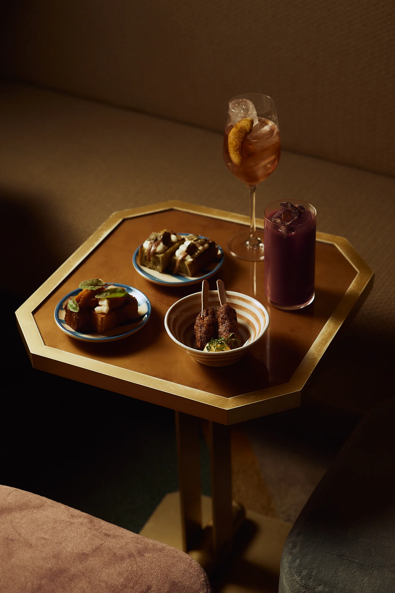 Octagonal table with two drinks and three plates of artisanal appetizers in a cozy setting.