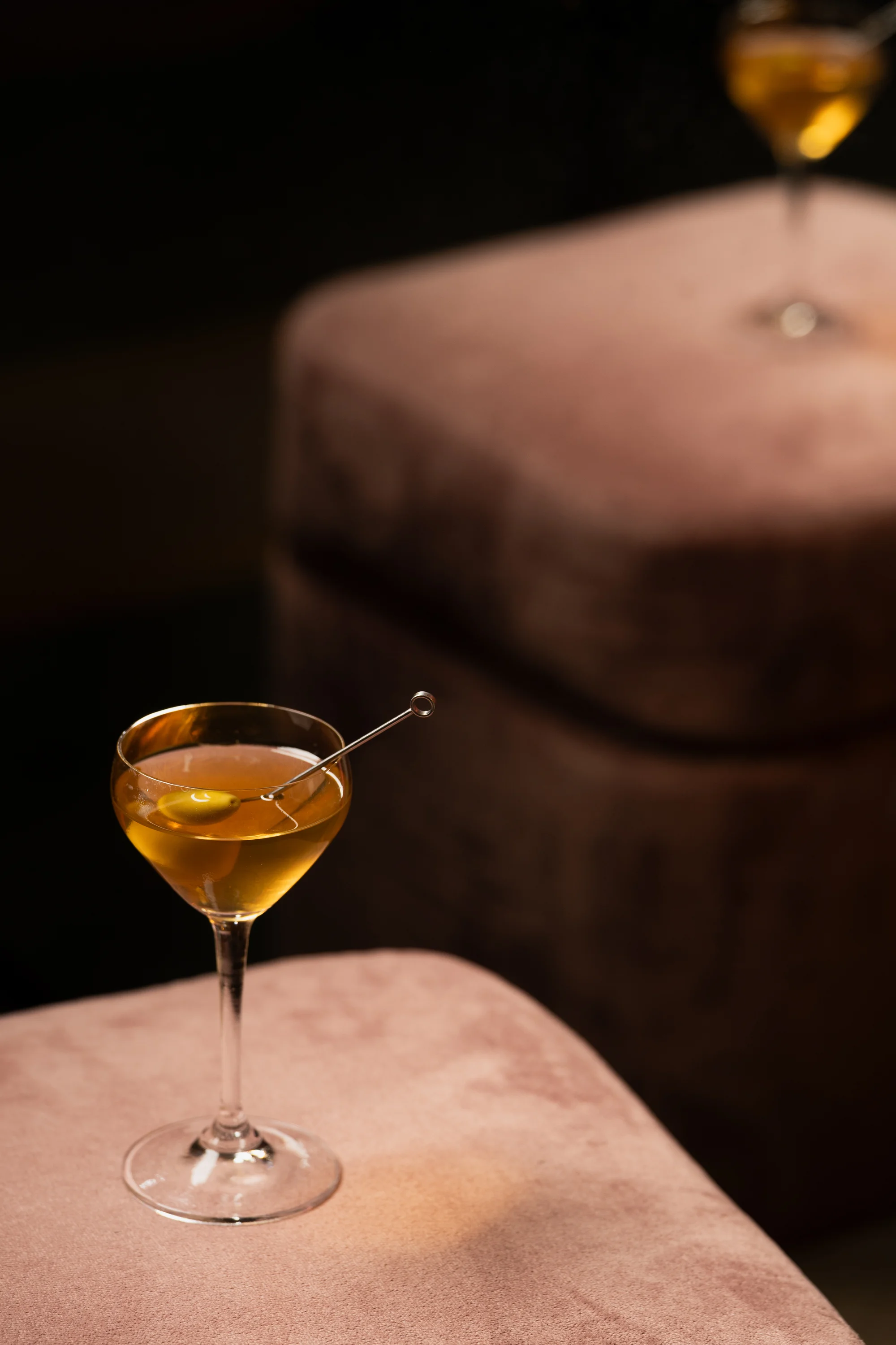 Cocktail glass with an olive garnish on a pink velvet surface in soft lighting.