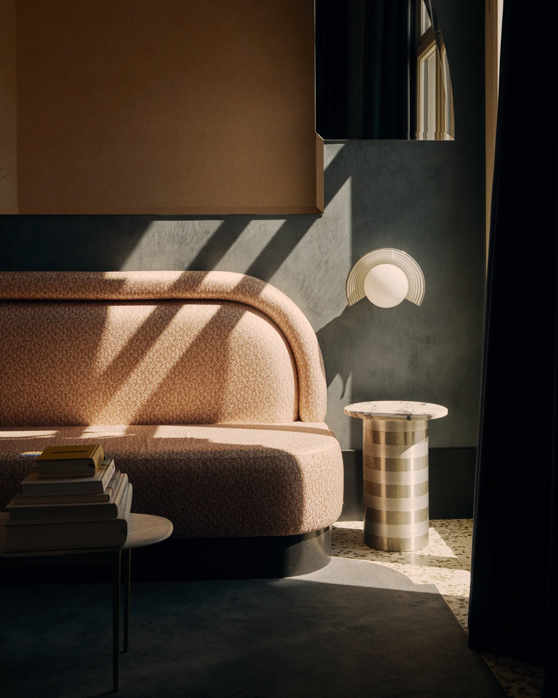 Curved beige sofa with stacked books on a side table and a striped pedestal table under sunlight.
