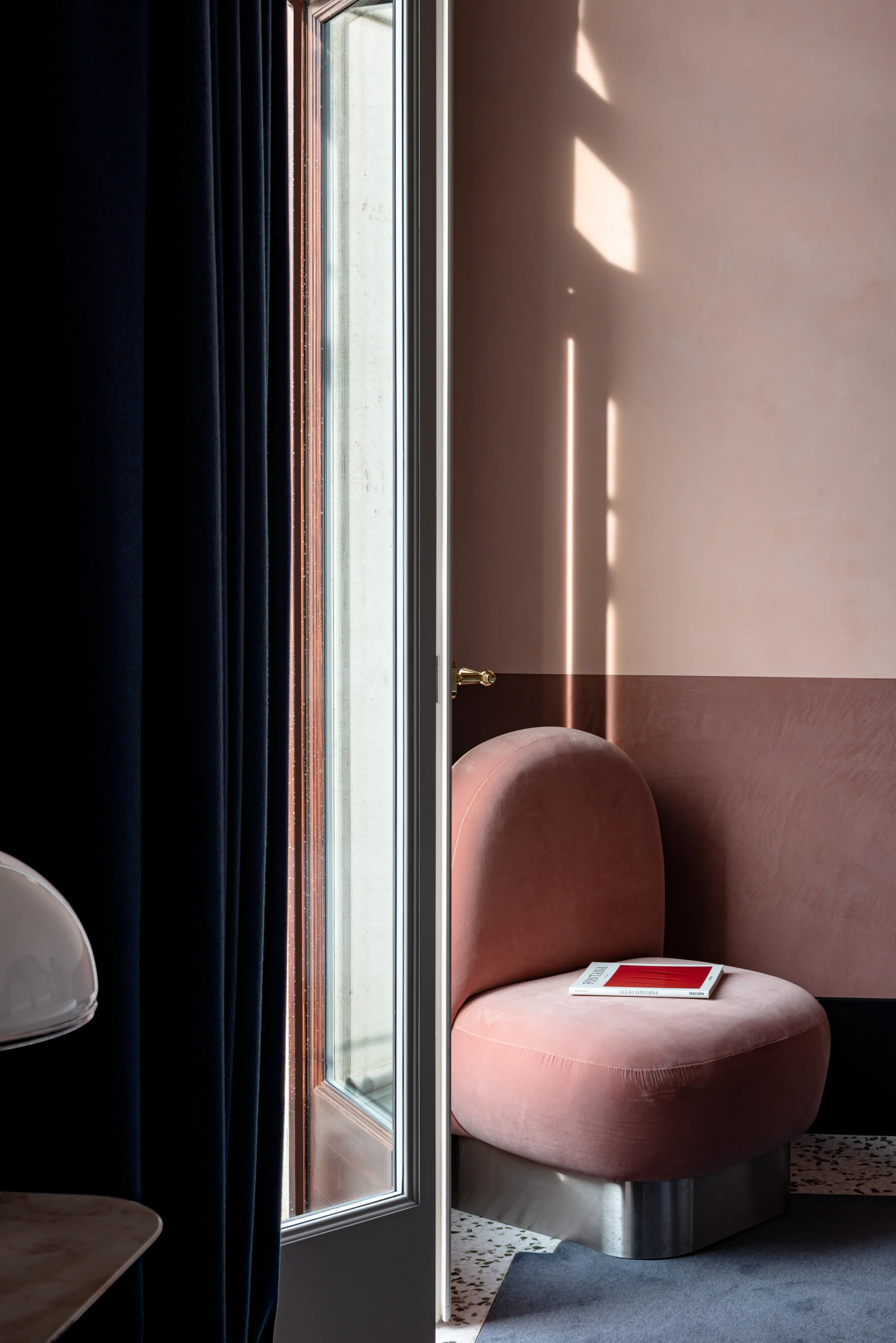 Silla de terciopelo rosa con un libro al lado de una puerta parcialmente abierta y cortinas oscuras.
