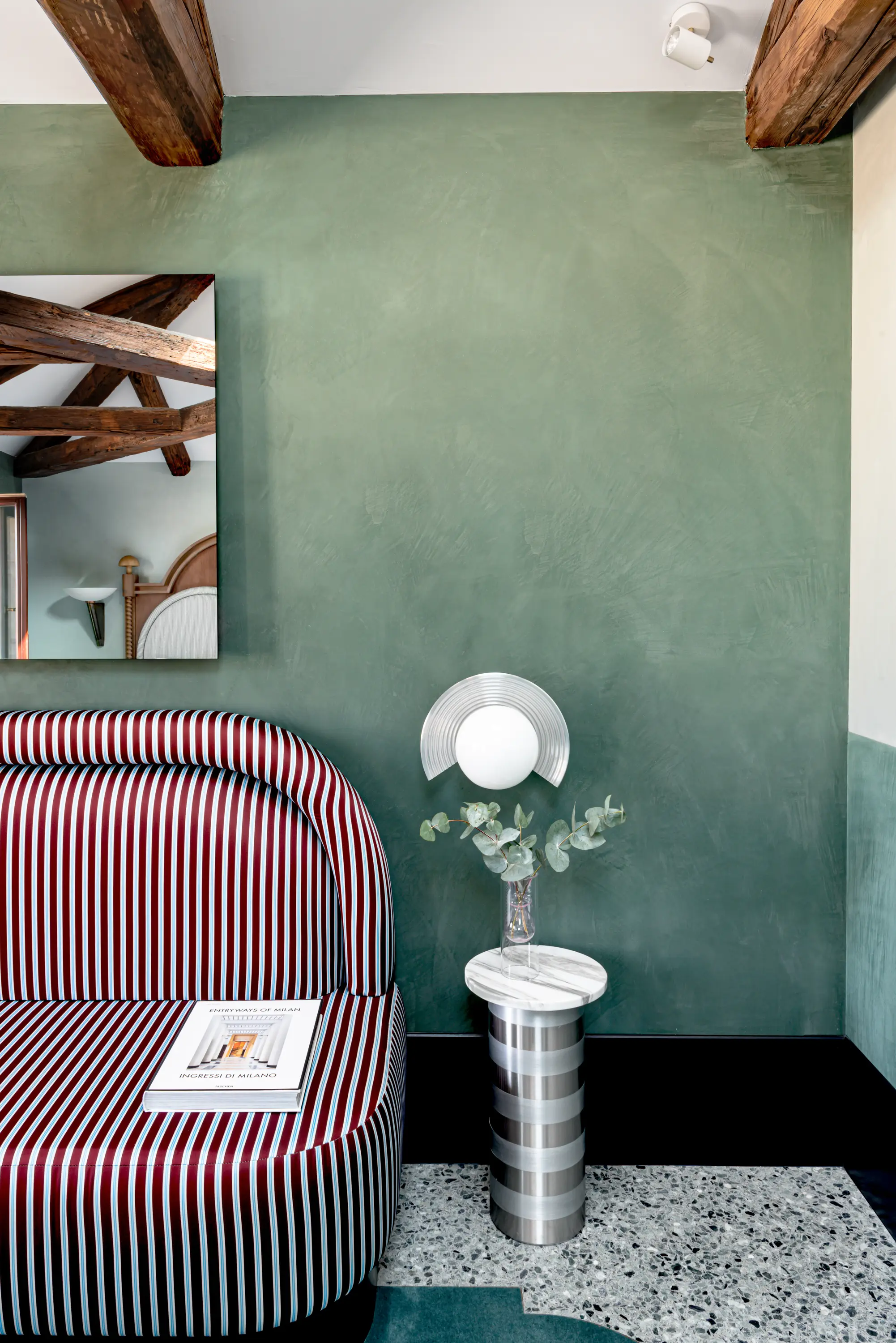 Sofá a rayas rojas y blancas junto a una mesa de metal y mármol con jarrón contra una pared verde texturizada.