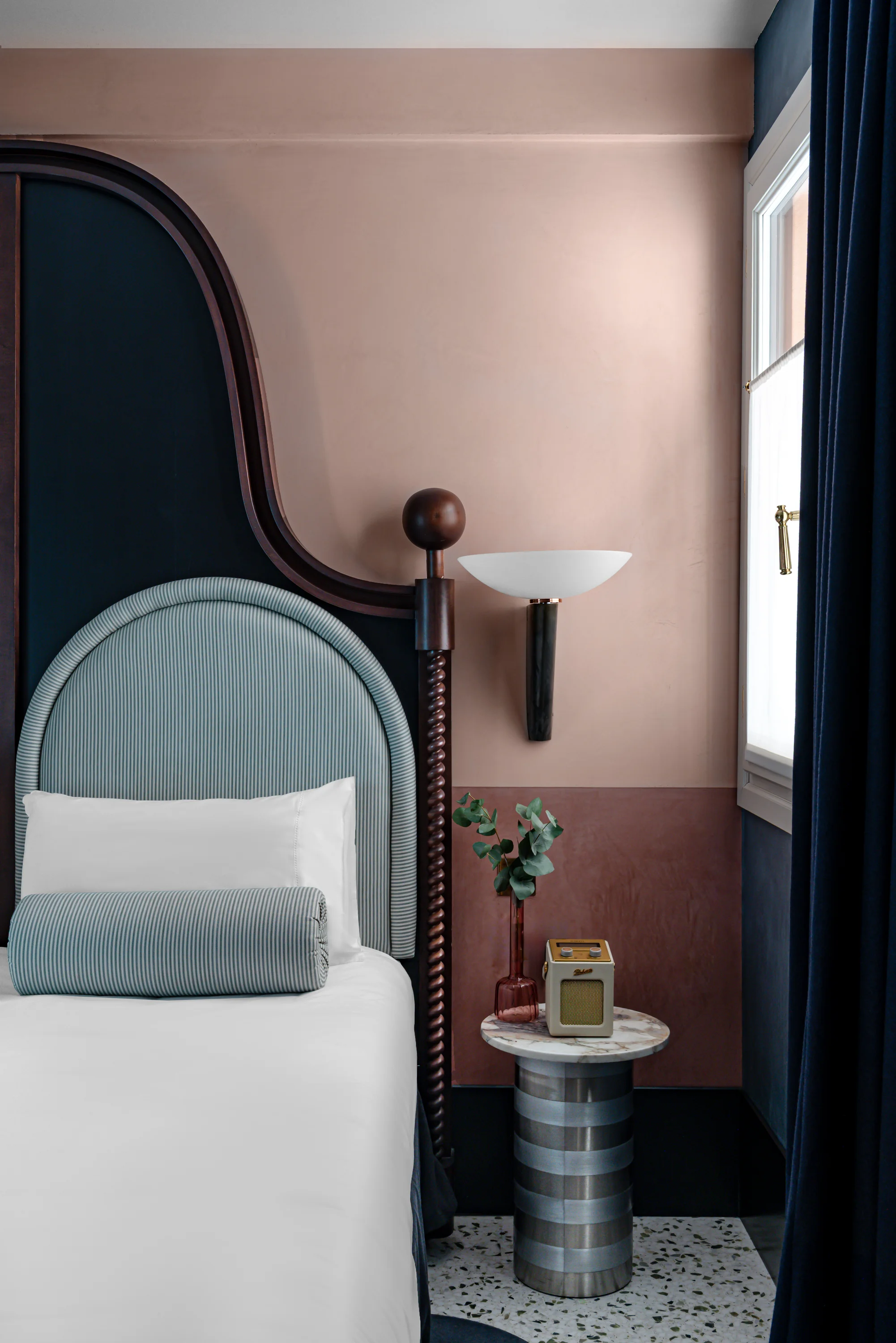 Esquina de dormitorio moderna con cabecera de rayas azules, ropa de cama blanca, mesa auxiliar de mármol y radio vintage.