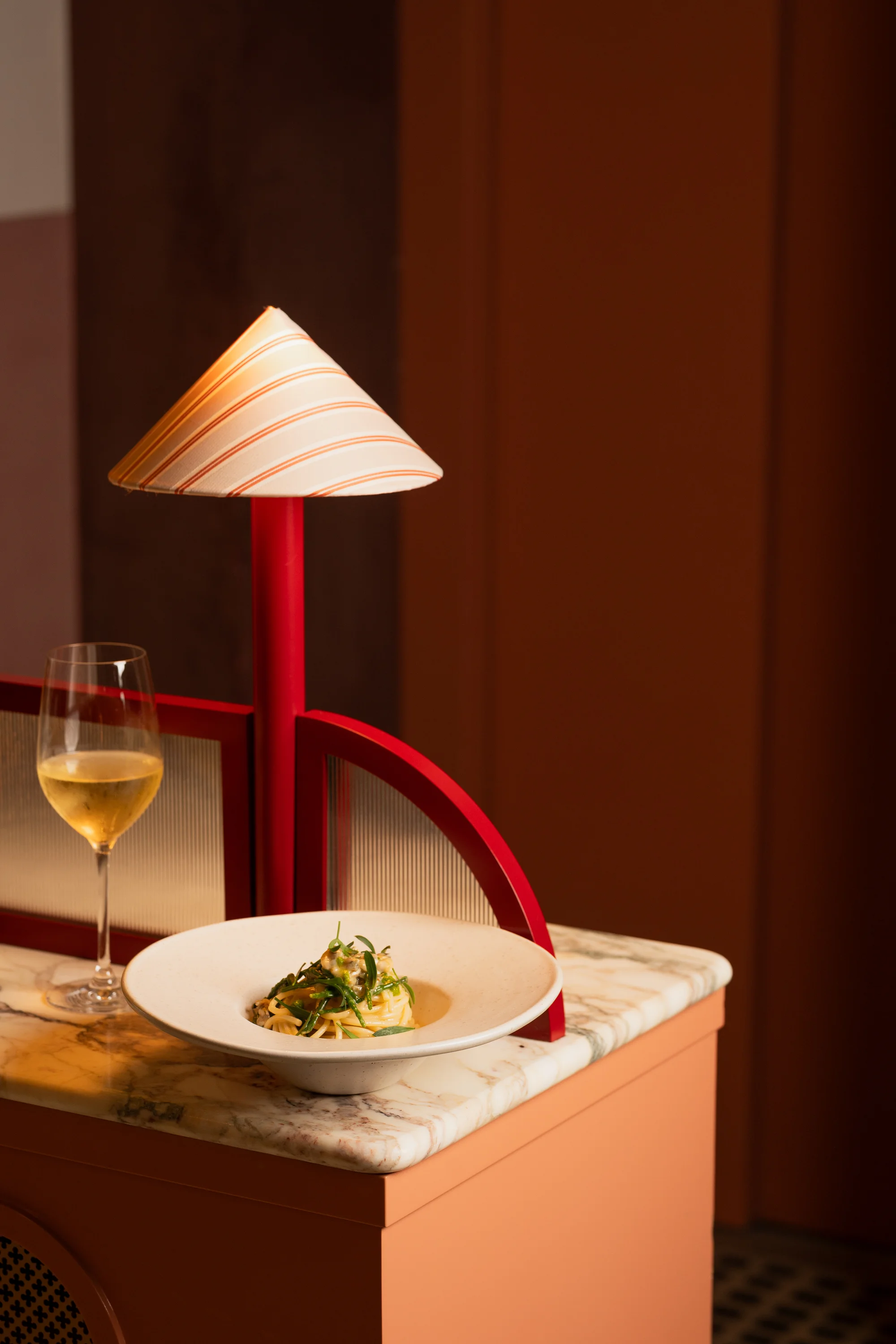 Cuenco blanco con pasta y hierbas sobre una tapa de mármol junto a una copa de vino blanco y una lámpara roja y beige.