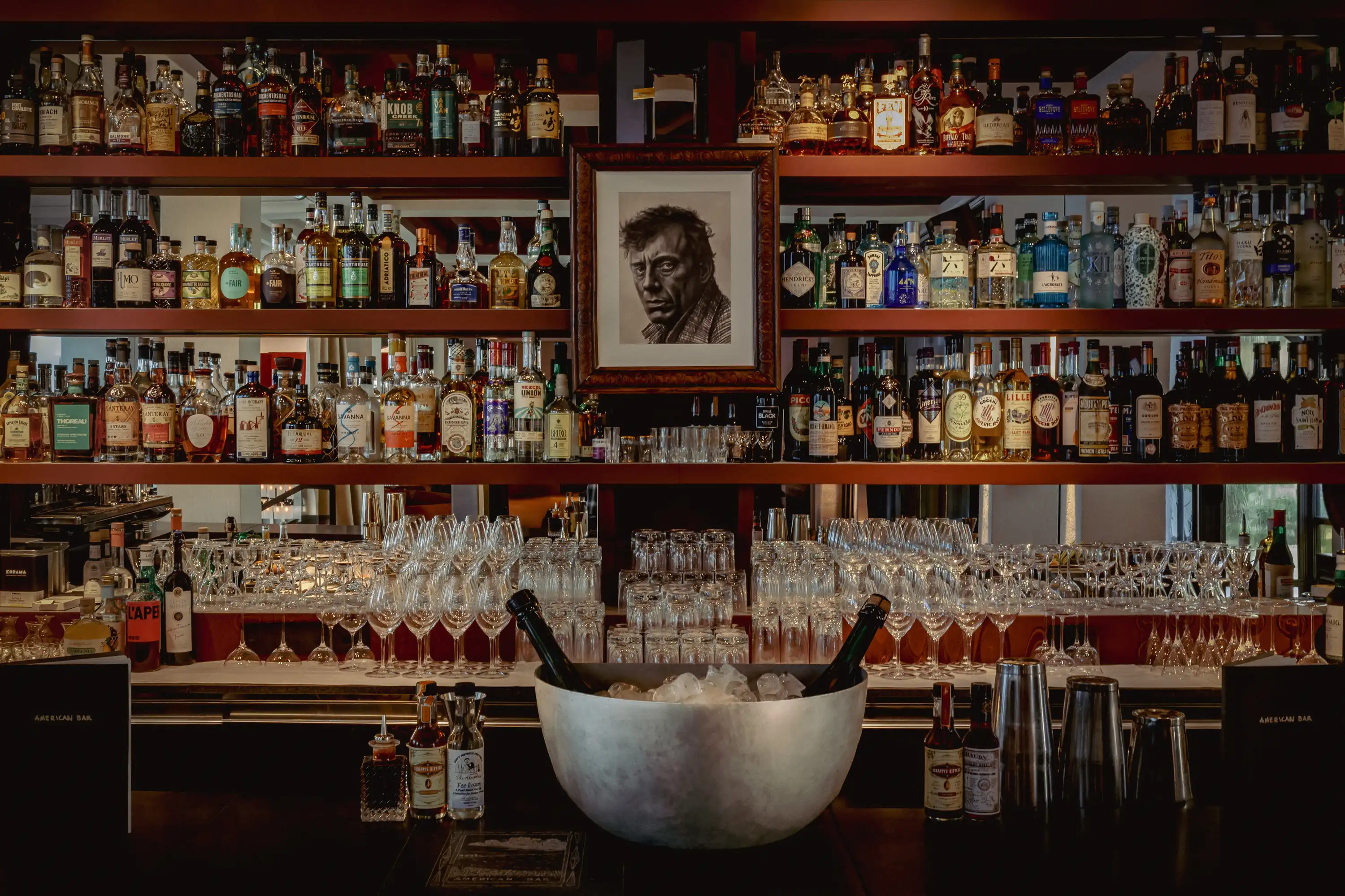 Bar counter with shelves of liquor bottles, stacks of glasses, and a large ice bucket with two bottles.
