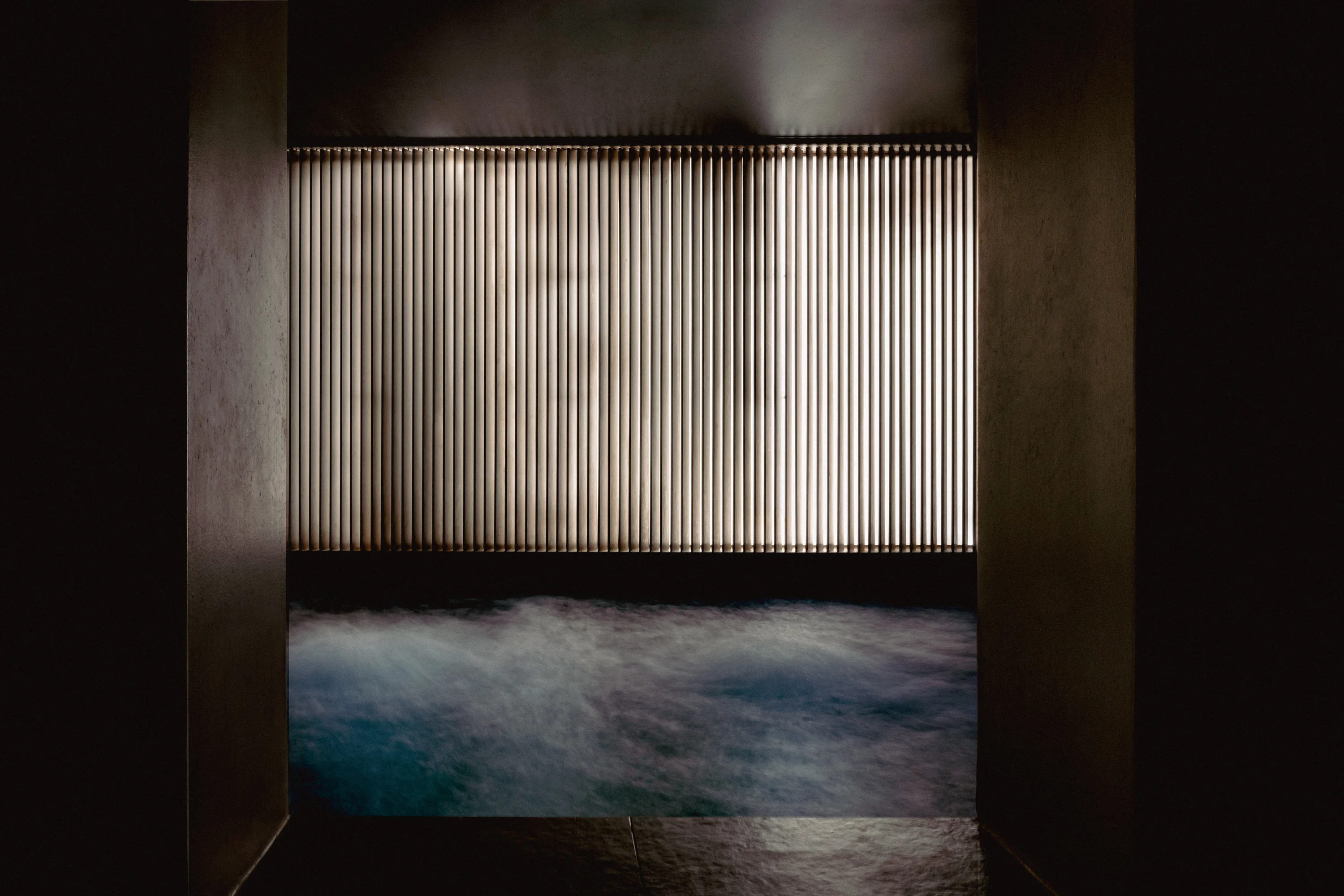 Dimly lit indoor pool with vertical blinds backlit behind steaming water.