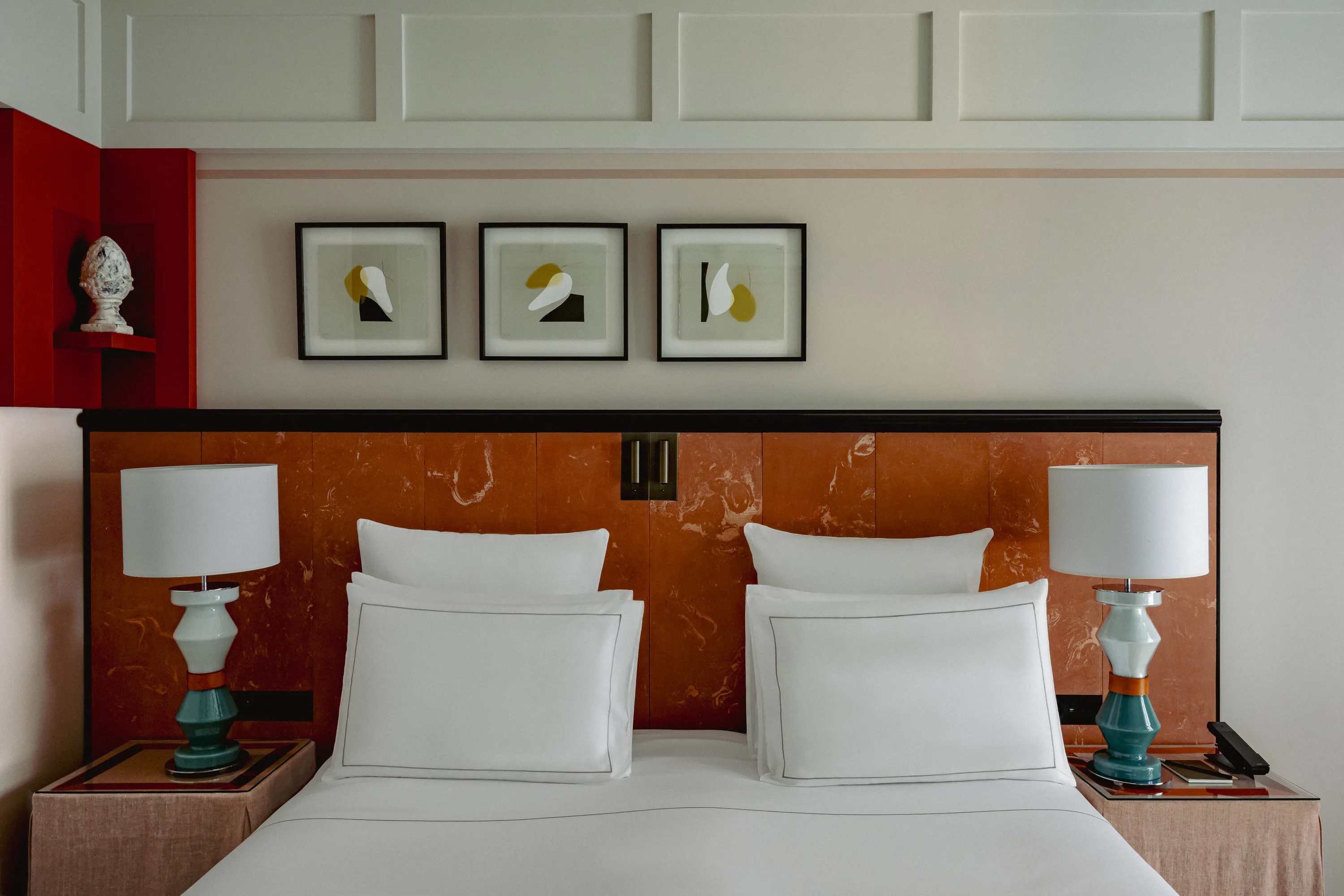 Modern bedroom with white pillows, teal lamps, and three abstract framed artworks above the bed.