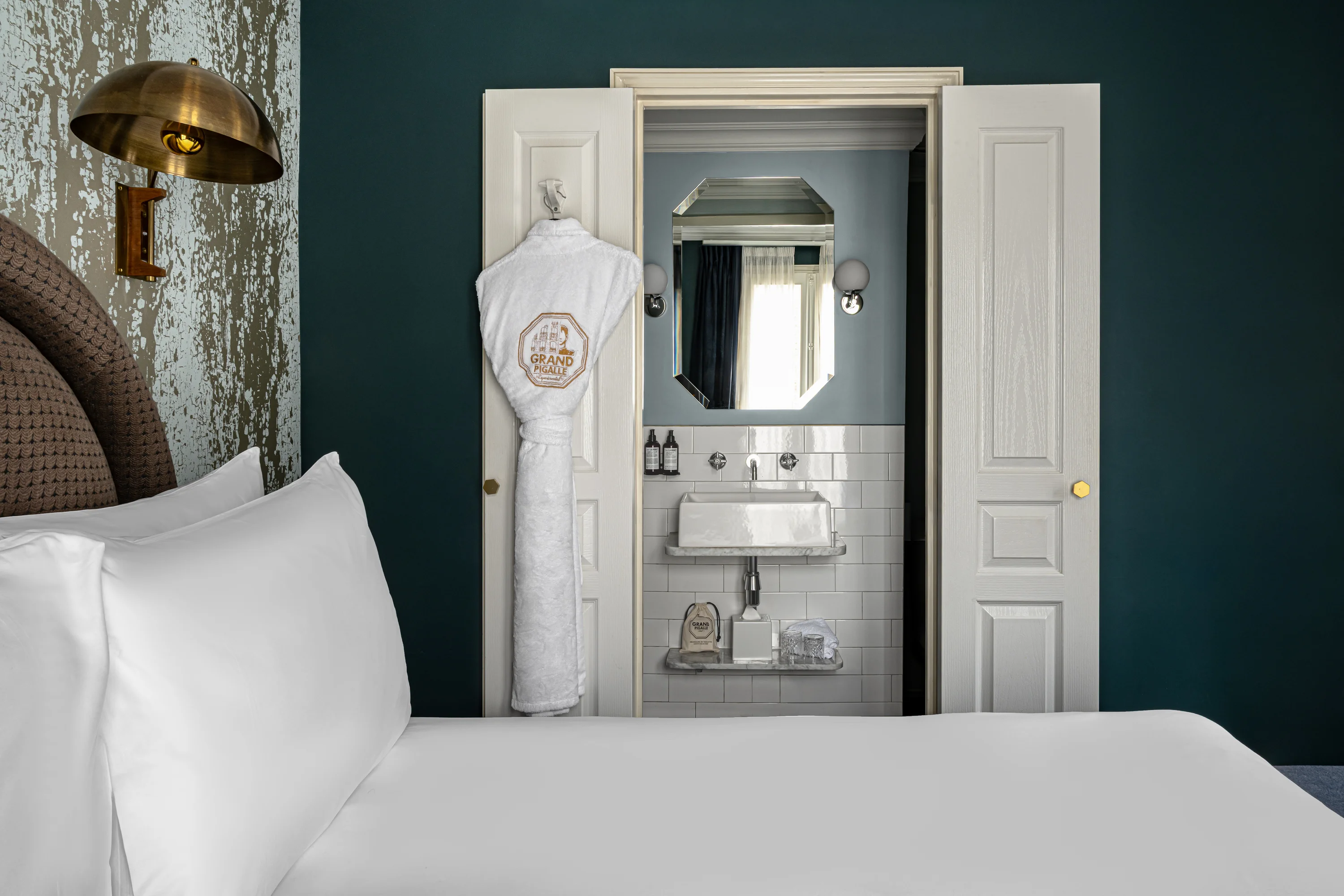Hotel room bed with white sheets, a robe hanging on door, and a bathroom sink with mirror beyond.