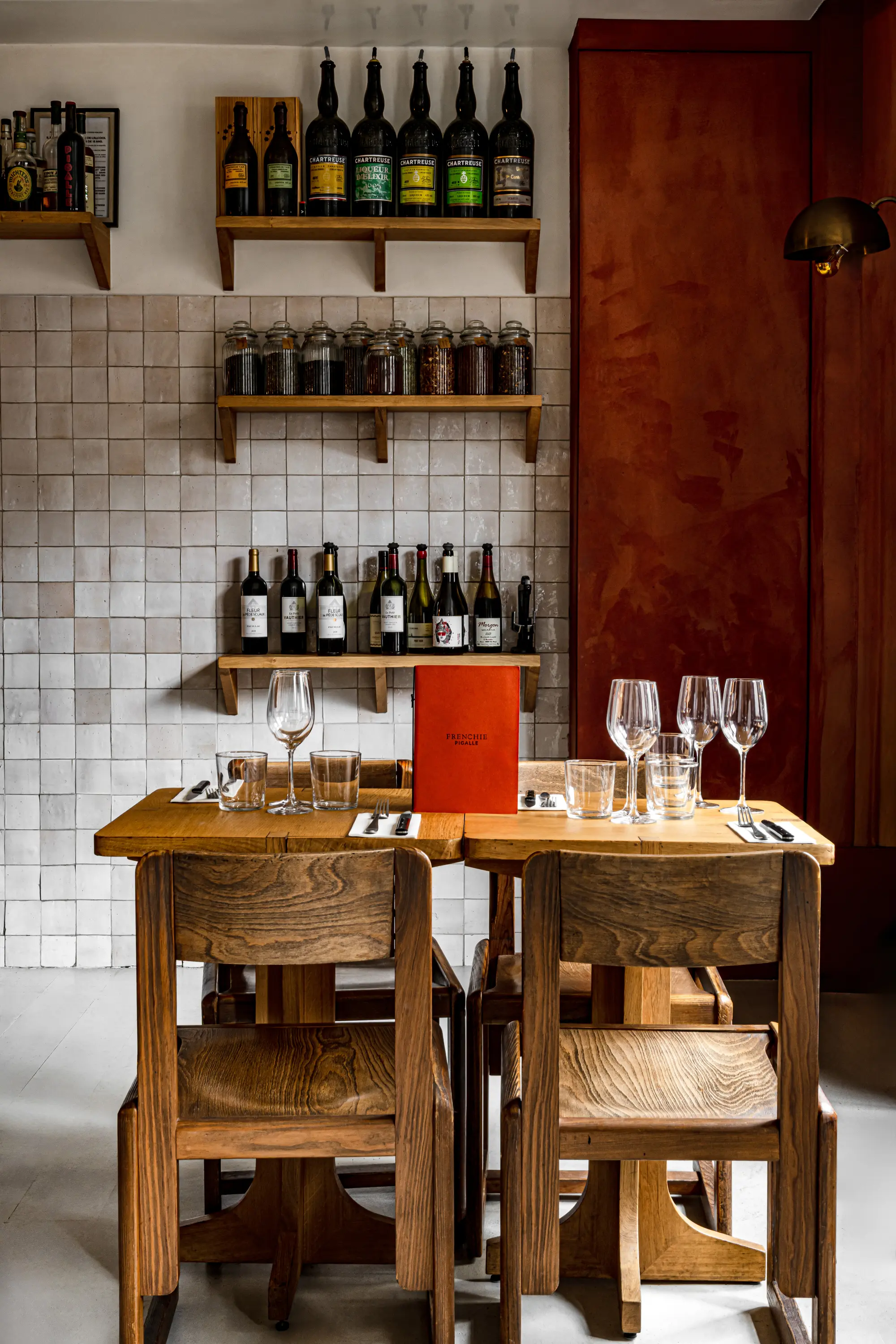 Wooden table with glasses, cutlery, and menu, with shelves of wine bottles and jars on a tiled wall.
