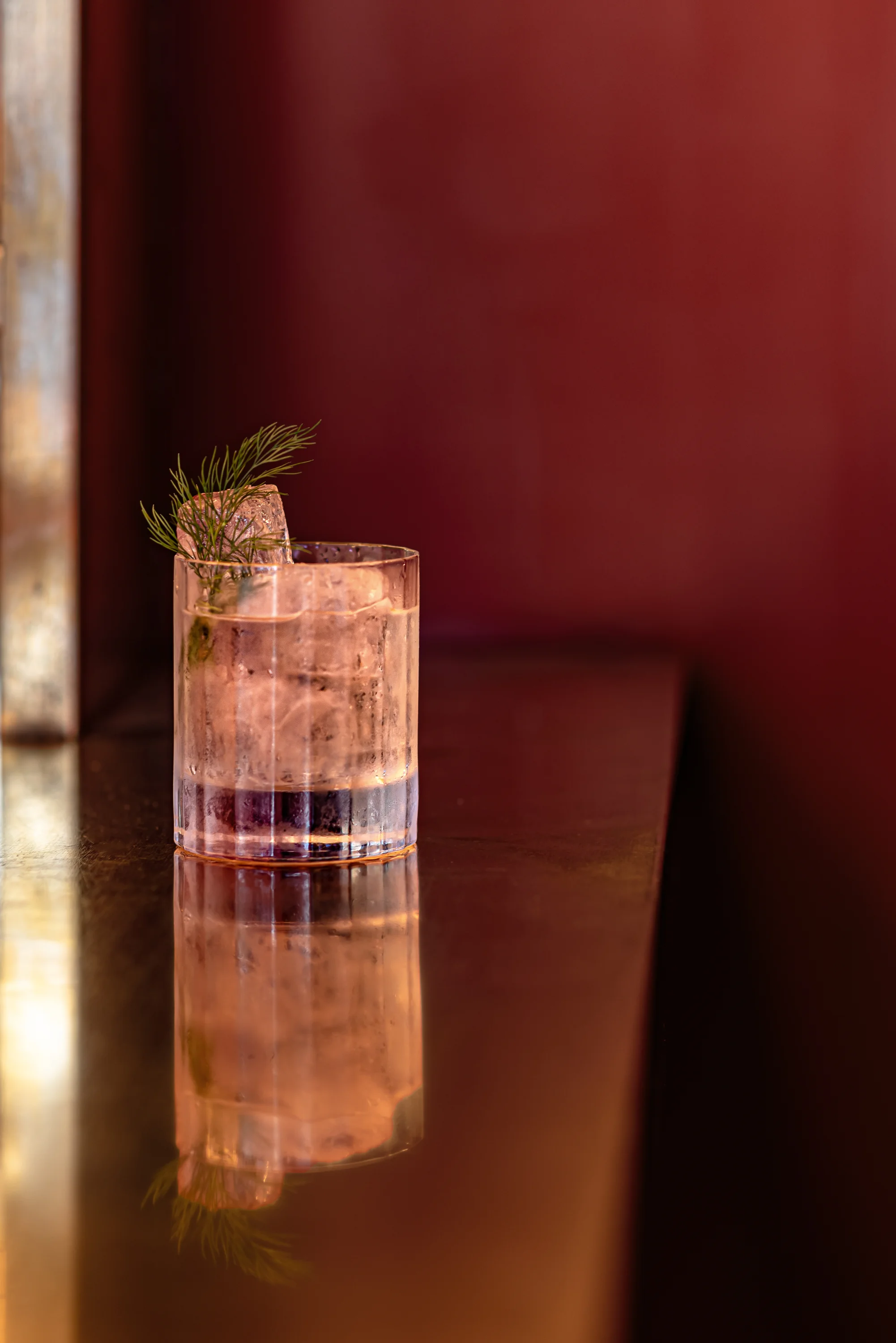 Clear cocktail with large ice cubes and dill garnish on reflective surface against a burgundy wall.
