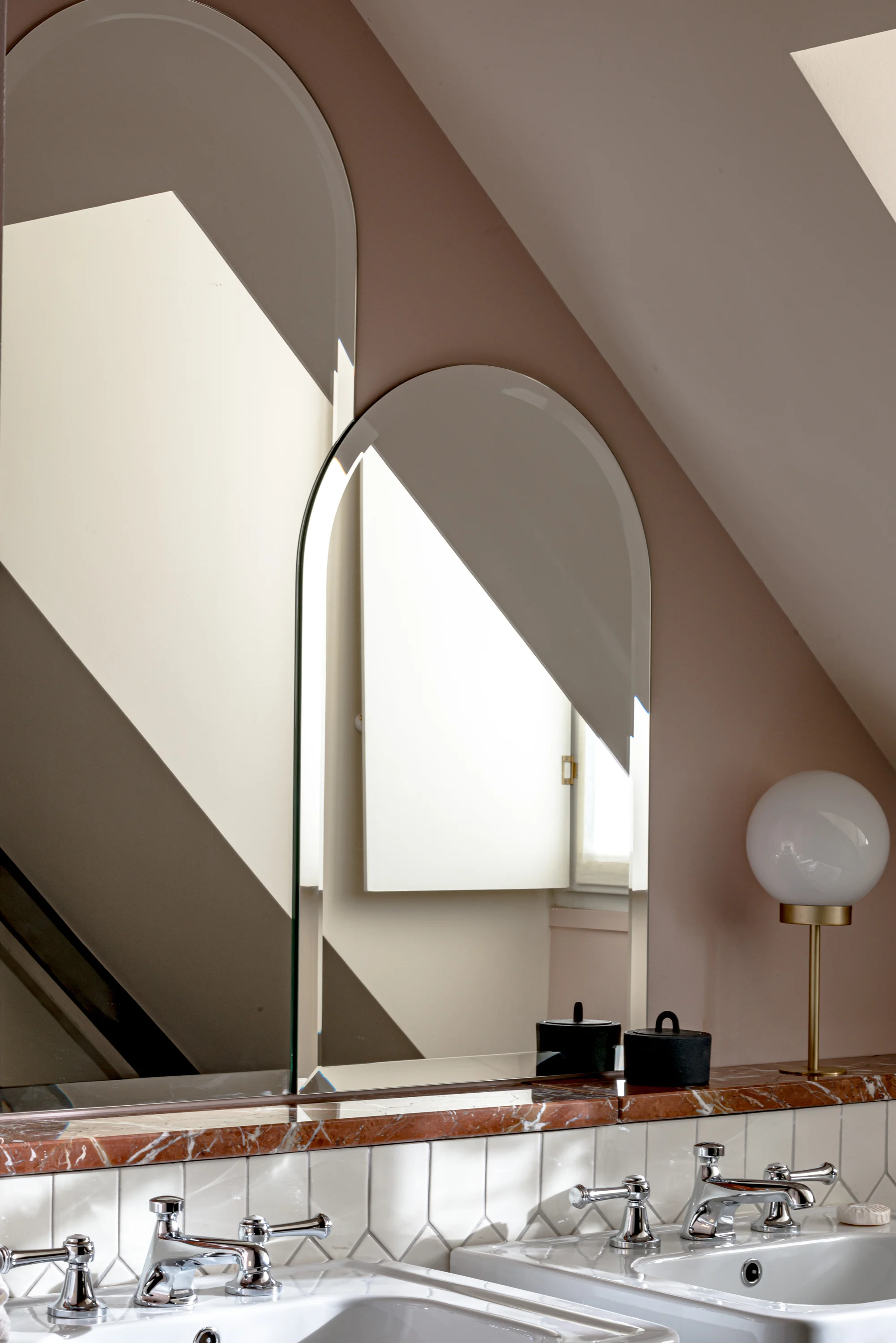 Bathroom countertop with two sinks, a round mirror, and a lamp with a white globe shade.