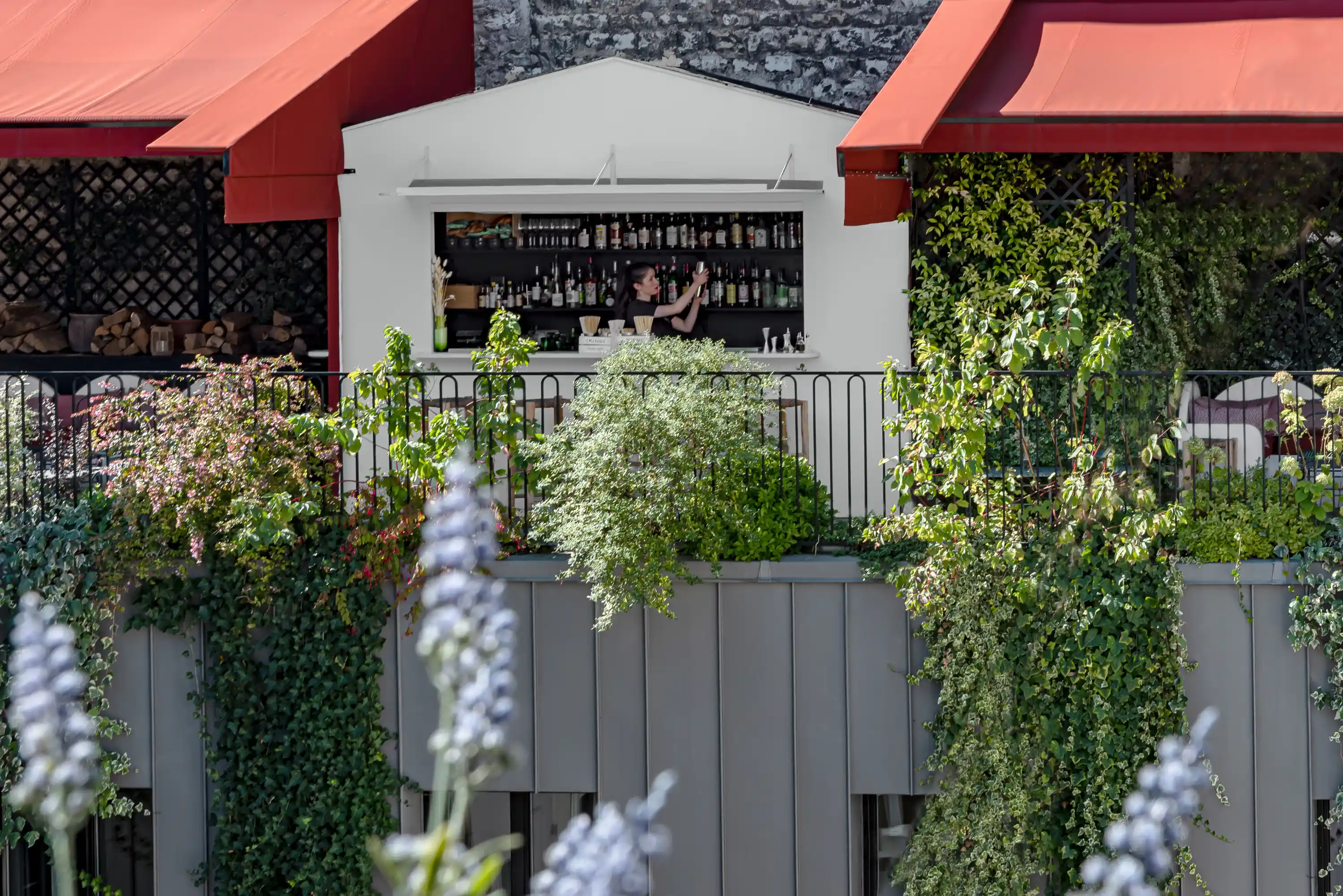 Woman bartender arranging bottles inside an outdoor white bar surrounded by green plants and red awnings.