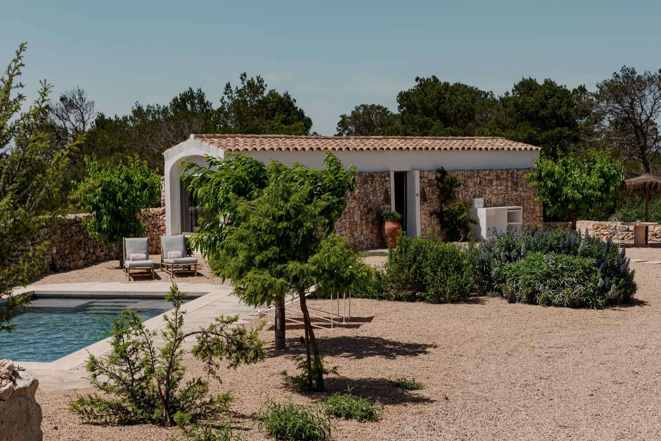 Stone house with tiled roof, pool, lounge chairs, and lush greenery in a sunny garden setting.