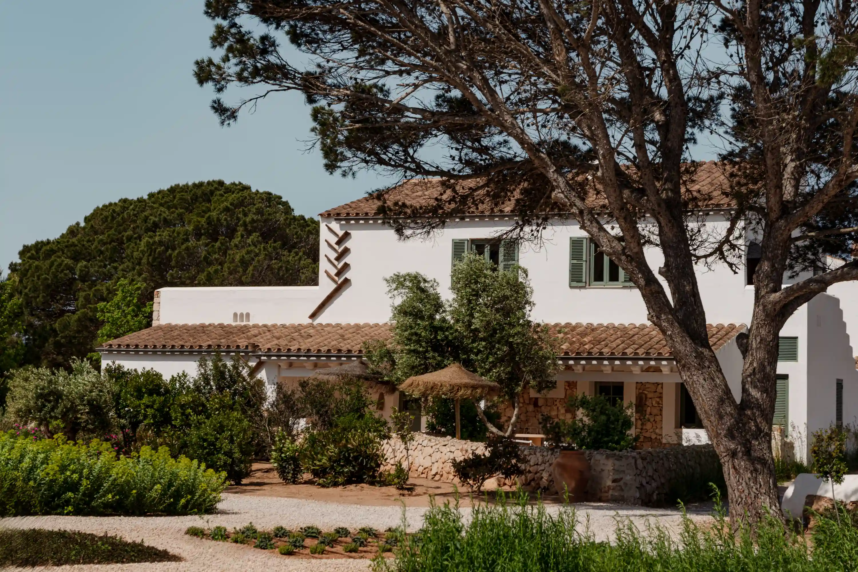 White Mediterranean house with terracotta roof, green shutters, surrounded by trees and garden paths.