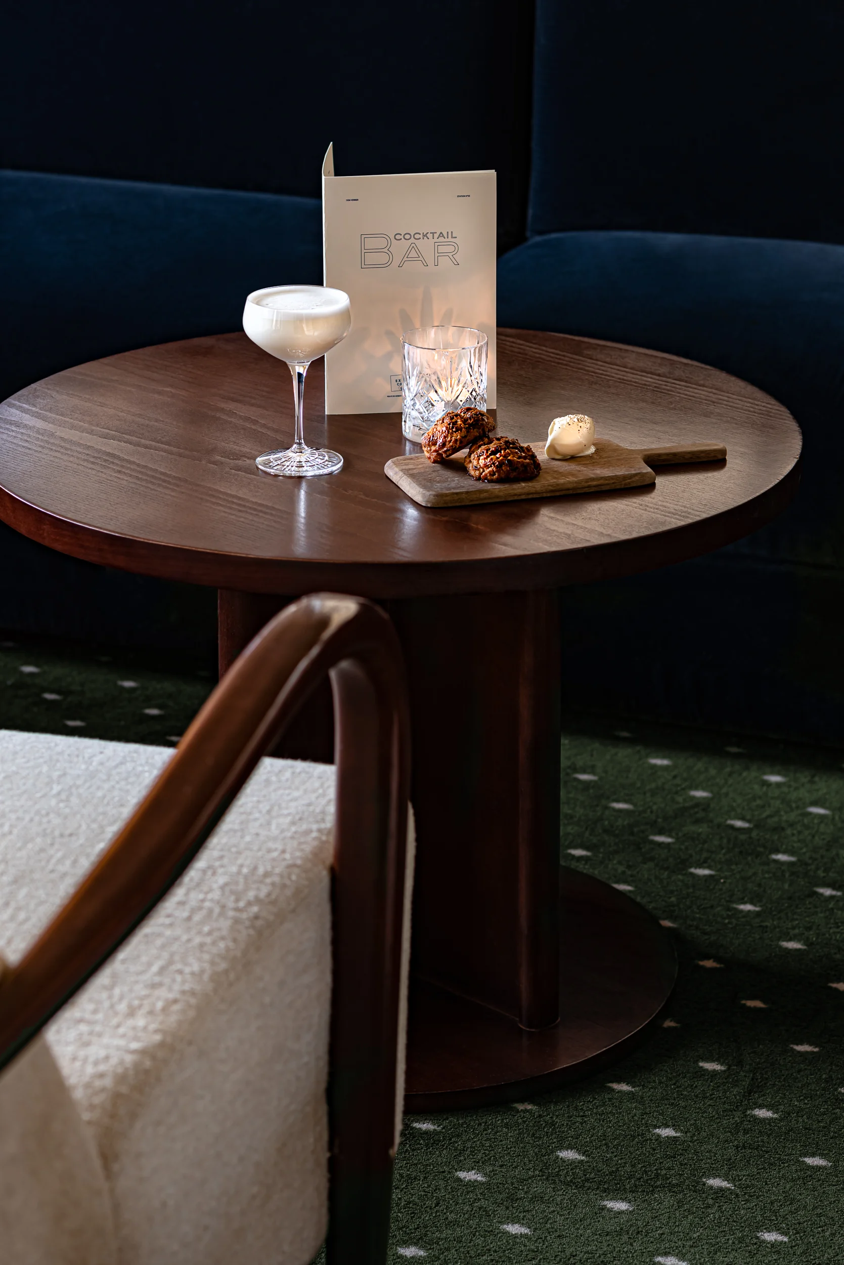 Cocktail glass, two cookies with cream on a cutting board, and a menu on a round wooden table.