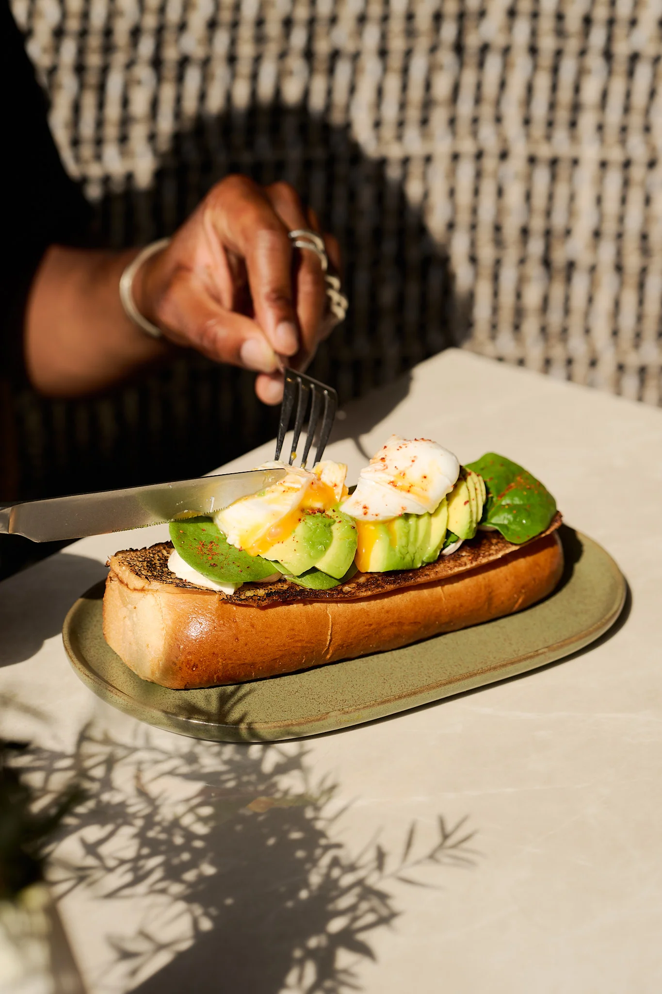Hand cutting into toast topped with sliced avocado, spinach, and a poached egg on a green plate.