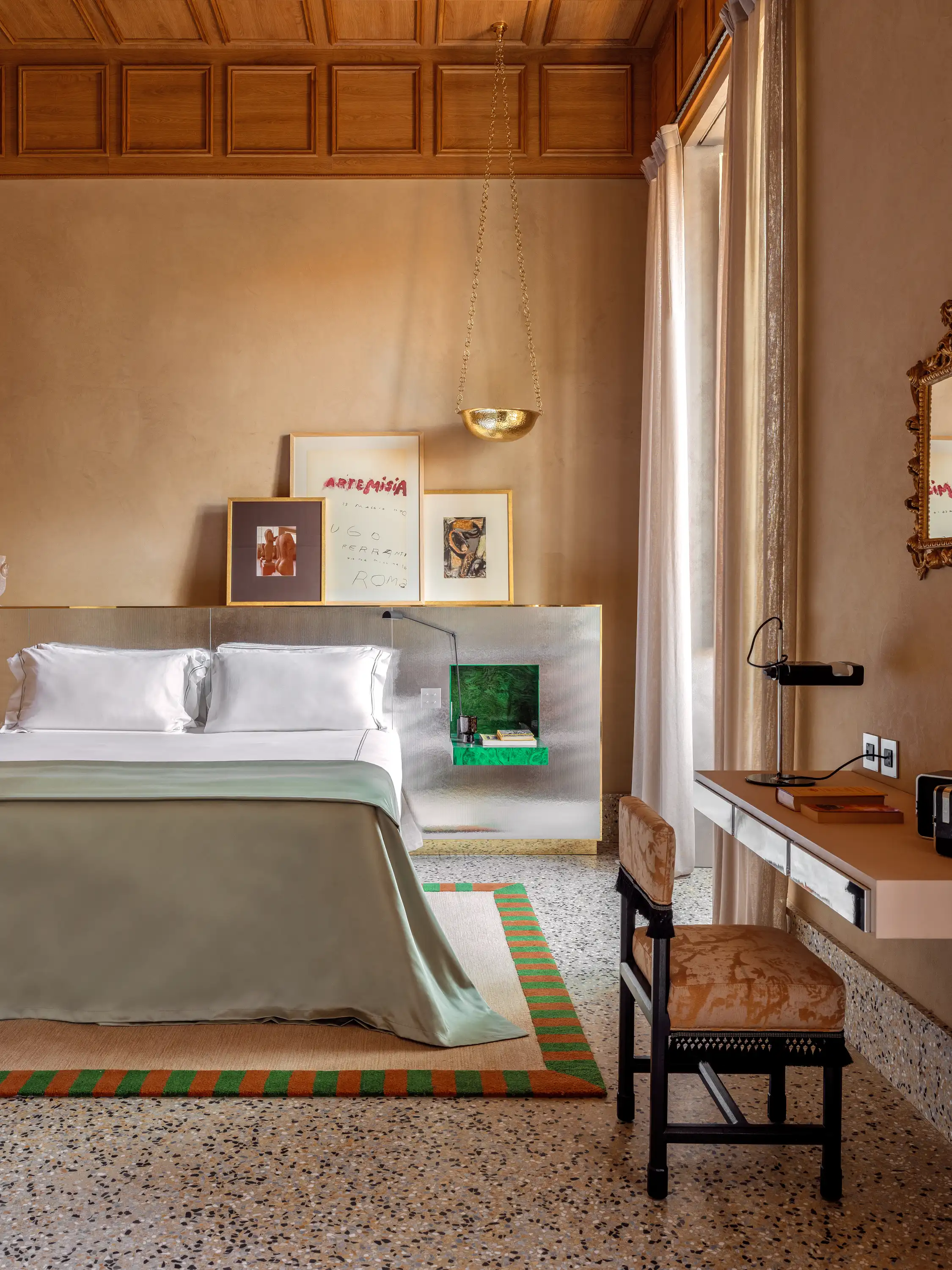 Minimalist bedroom with white bed, green and orange rug, gold hanging lamp, and elegant workspace.