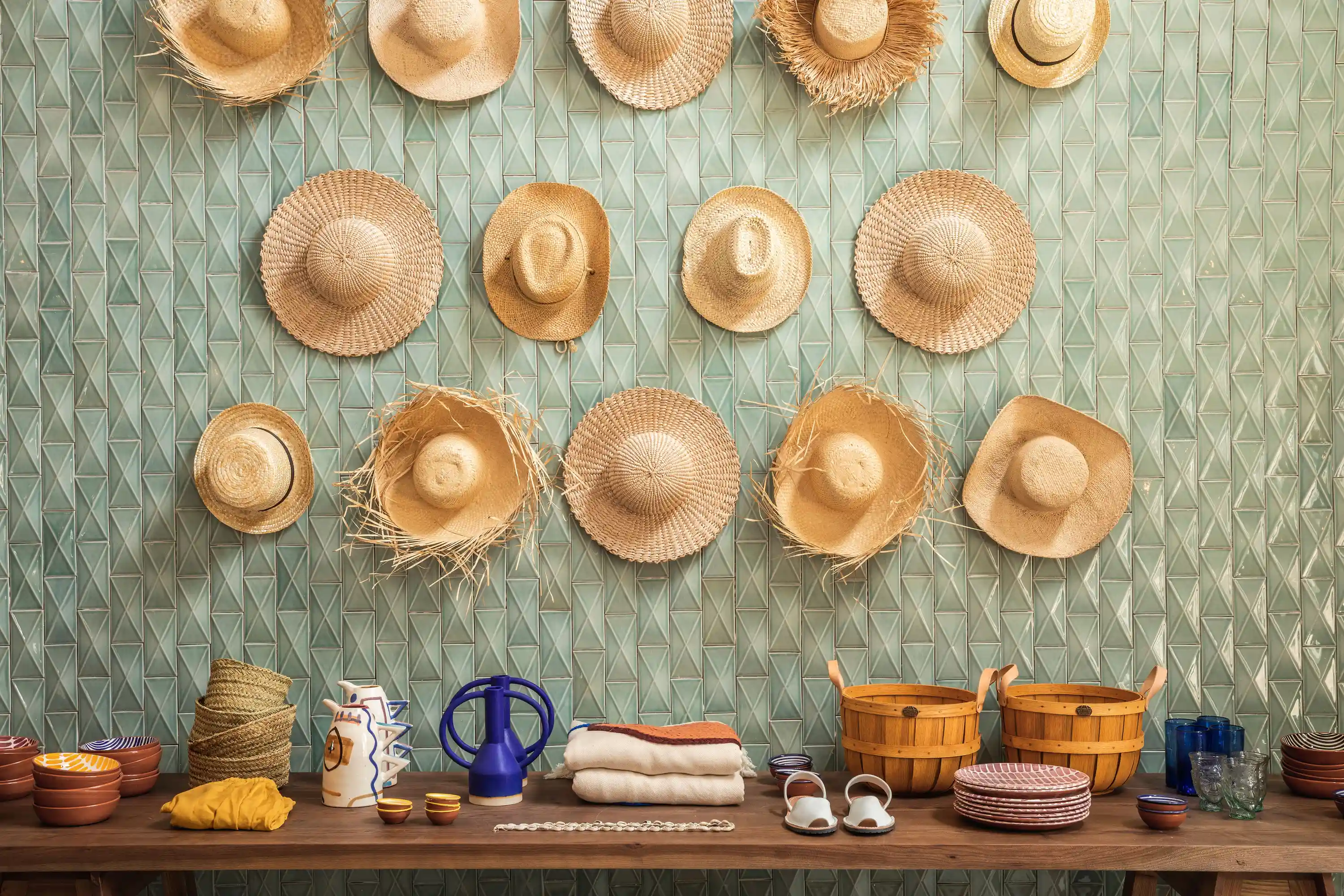 Various straw hats hang on a green tiled wall above a wooden table with ceramics and baskets.