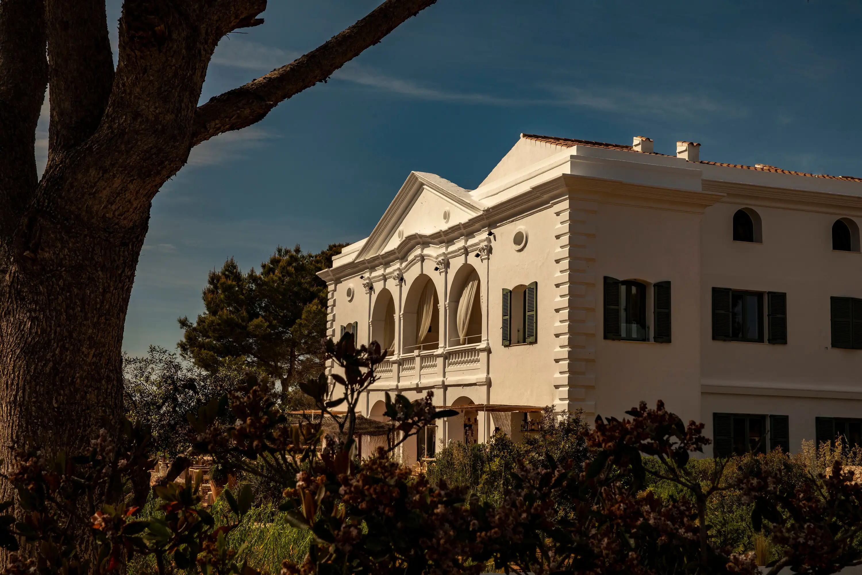 Sunlit white villa with arched balcony and dark windows among trees and bushes under blue sky.