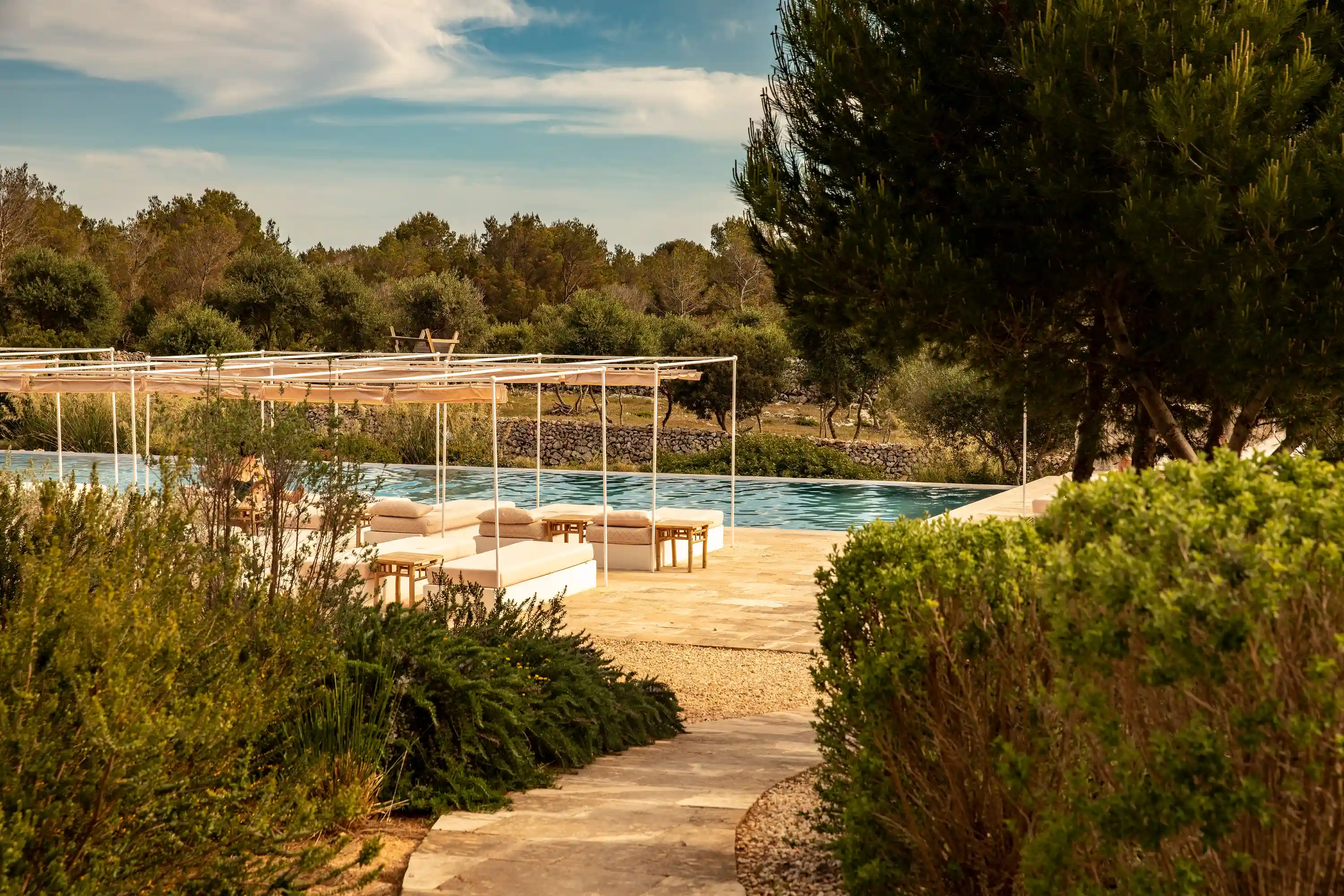 Outdoor pool area with white canopy loungers surrounded by dense greenery and trees under blue sky.