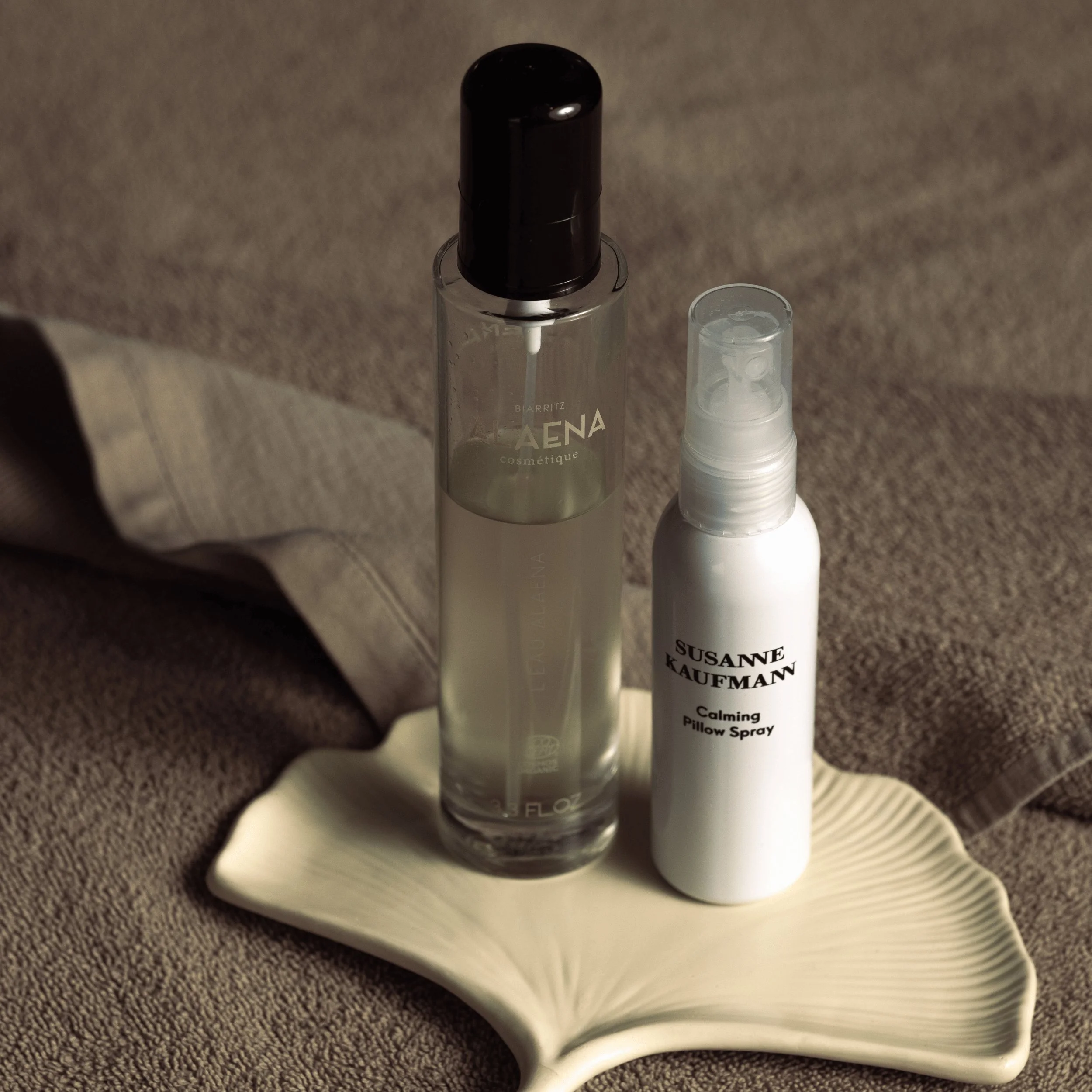 Due flaconi spray cosmetici posizionati su un piatto di foglie di ceramica beige su una morbida superficie di tessuto marrone.