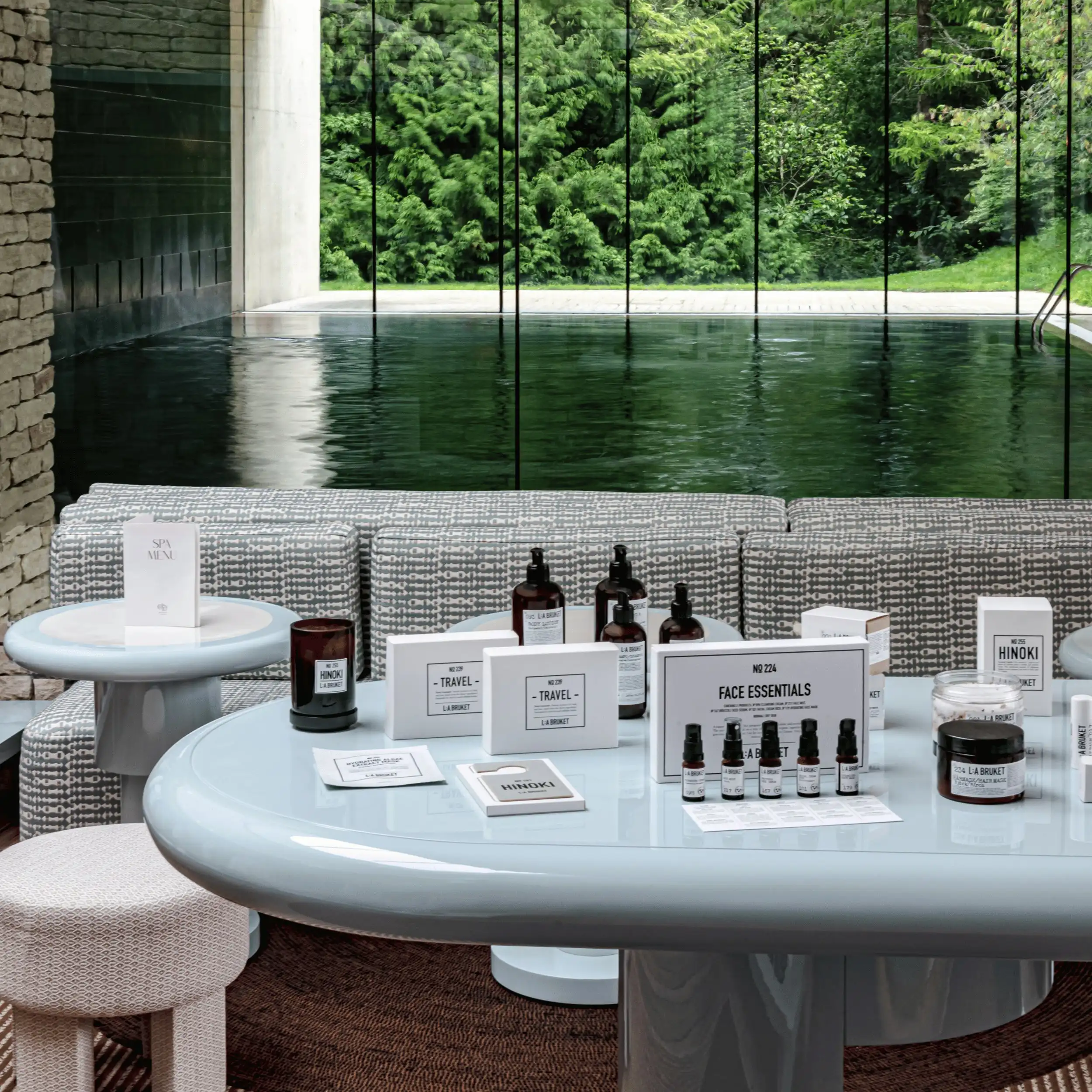 Prodotti per il benessere della spa esposti su un tavolo moderno con piscina e alberi verdi visibili attraverso il vetro.