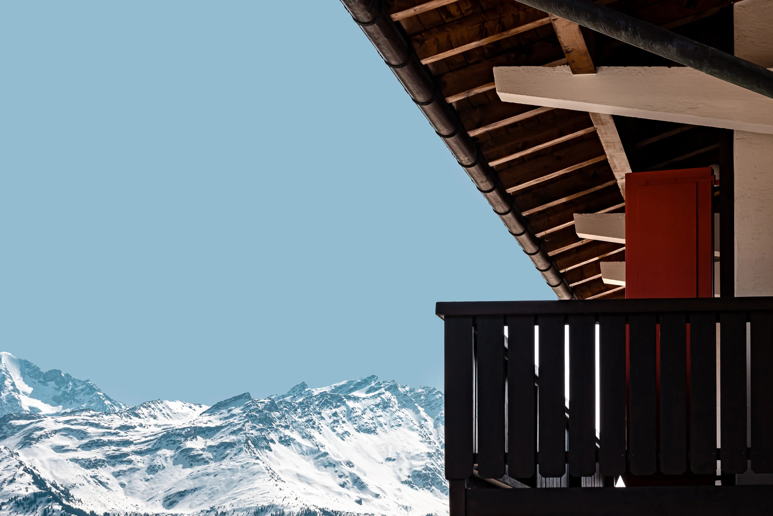 Balcony with wooden railing and red shutter overlooks snow-covered mountain peaks under clear blue sky.